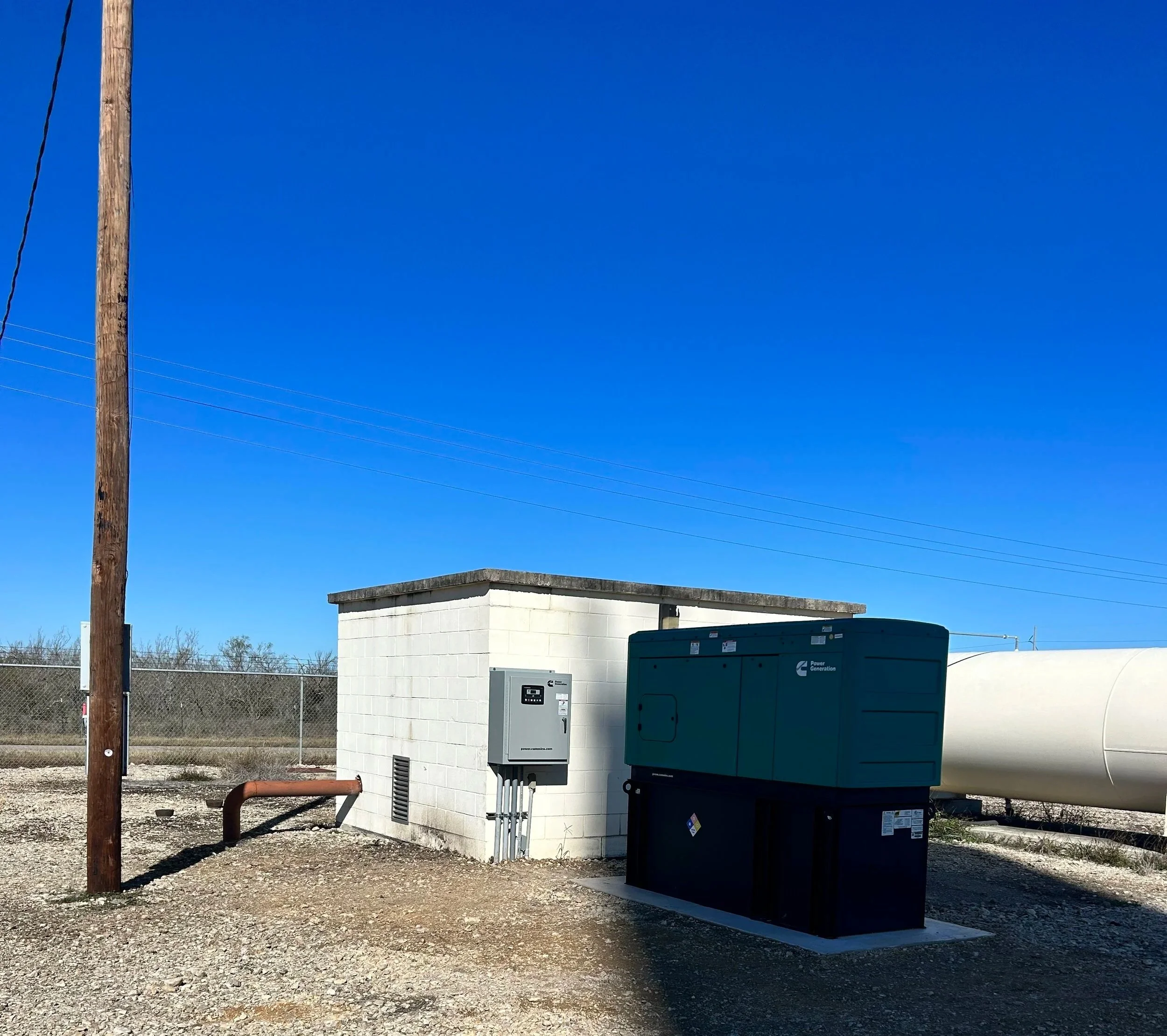 An outdoor scene with a utility pole on the left, a small white brick building with electrical and power equipment, a chain-link fence in the background, and a clear blue sky overhead.