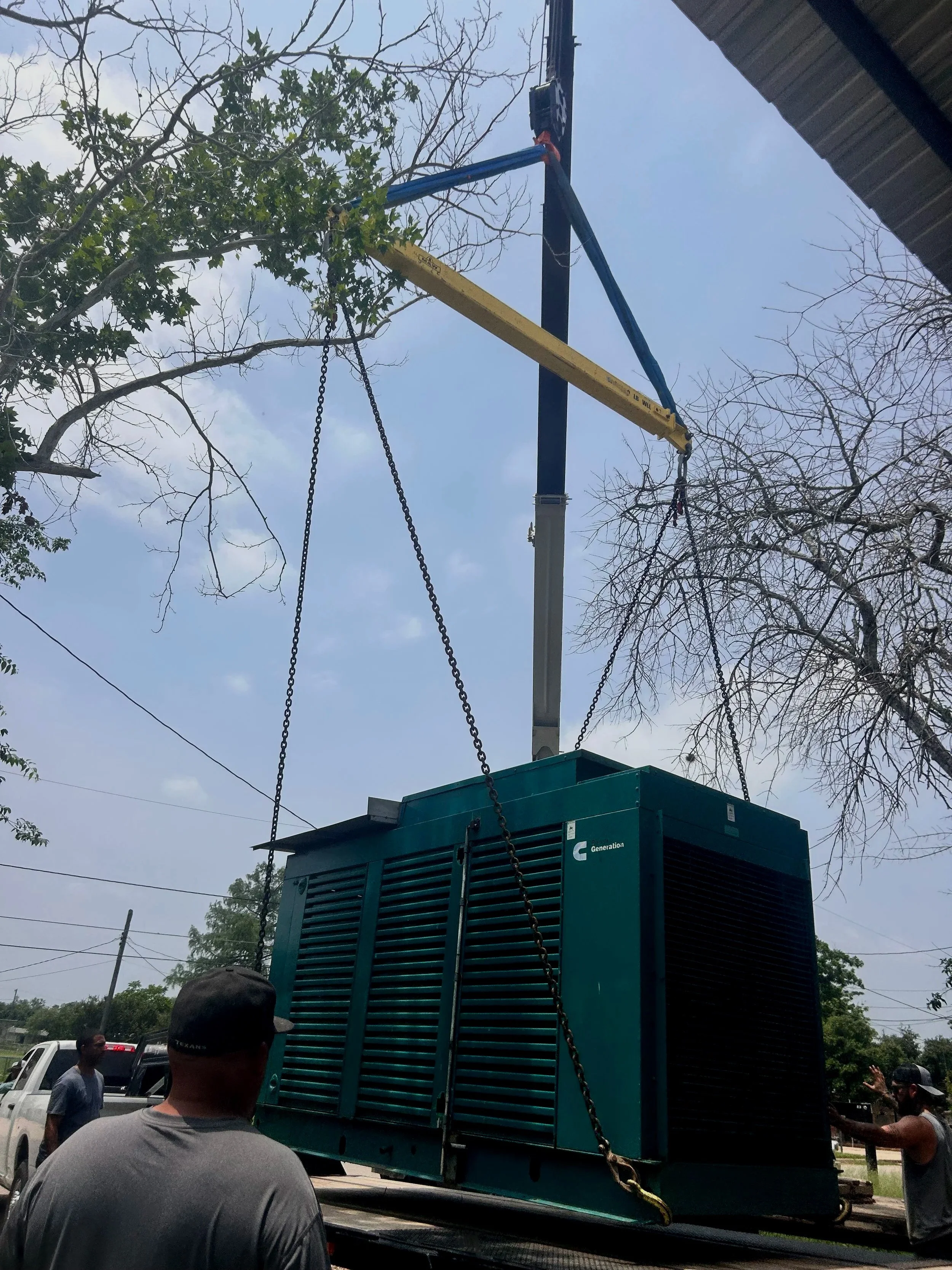 A portable generator being transported on a flatbed truck, with some people standing around and observing. There are trees and wires visible in the background.