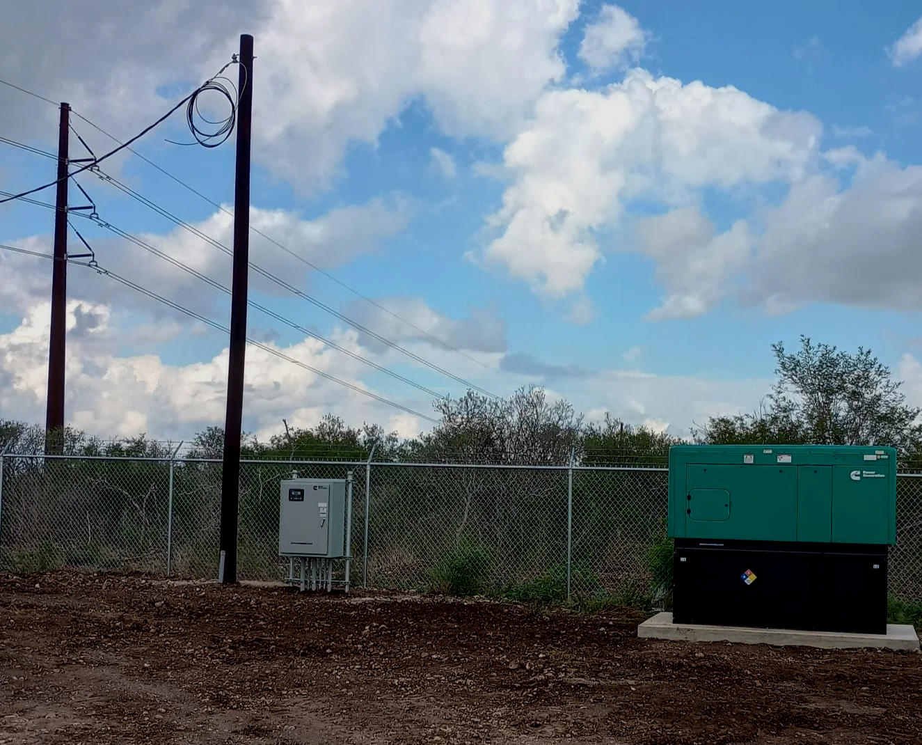 Electrical equipment and power lines outside, with a chain-link fence, trees, and a partly cloudy sky in the background.