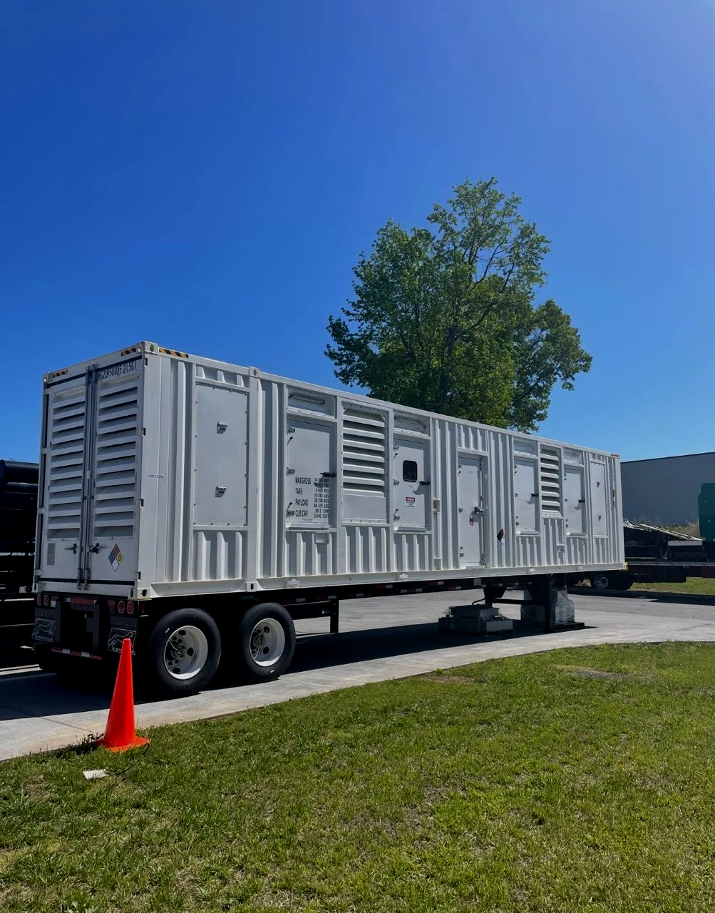White mobile generator trailer on a paved surface with a green grassy area, orange traffic cone, blue sky, and a tall green tree in the background.