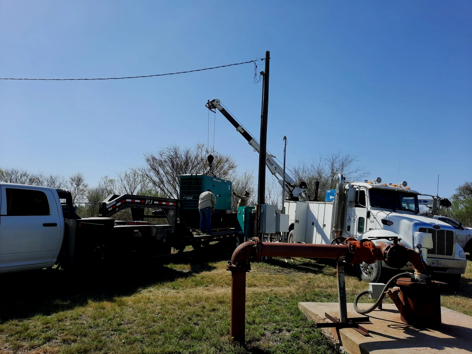 Workers operating utility equipment at a worksite with trucks, machinery, and a power pole against a clear blue sky.