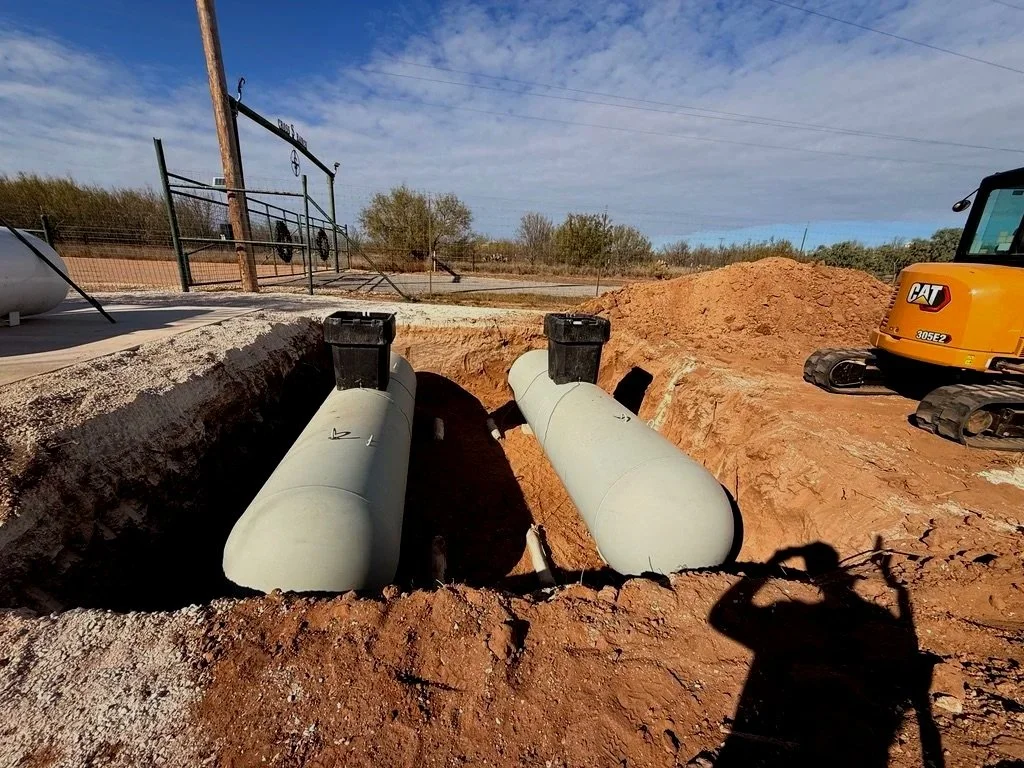 Underground pipeline installation at a construction site. Two large, light grey pipes are partially buried in a trench with a yellow excavator nearby. The site is outdoors under a partly cloudy sky.