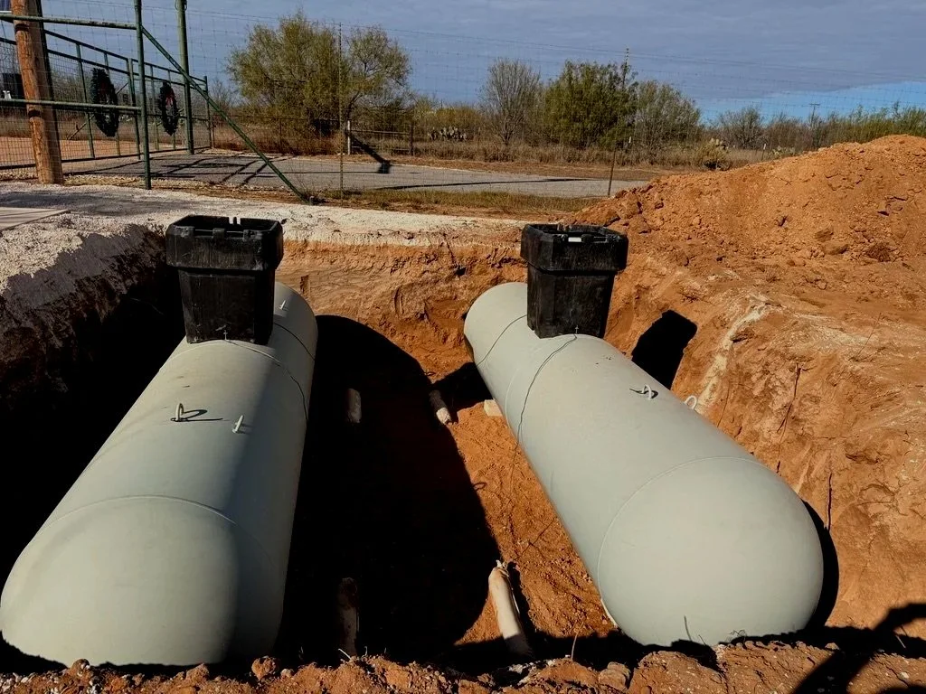 Two large underground pipes with black access boxes on top, buried in a construction site with dirt and a fenced outdoor area in the background.