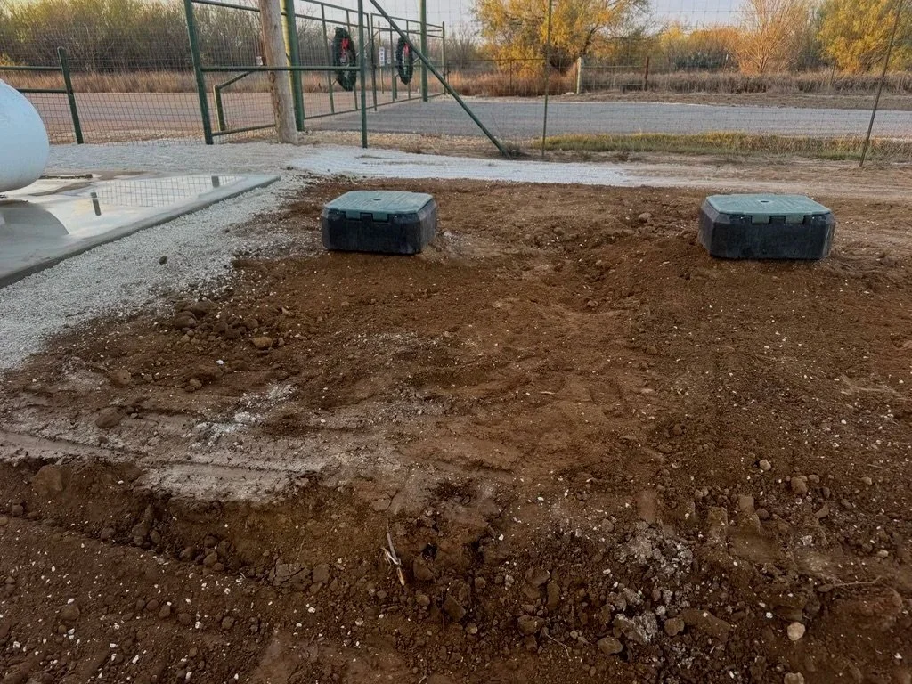 Construction site with dug-up dirt, two black plastic containers, a chain-link fence, and an open gate, set against a backdrop of trees and an overcast sky.
