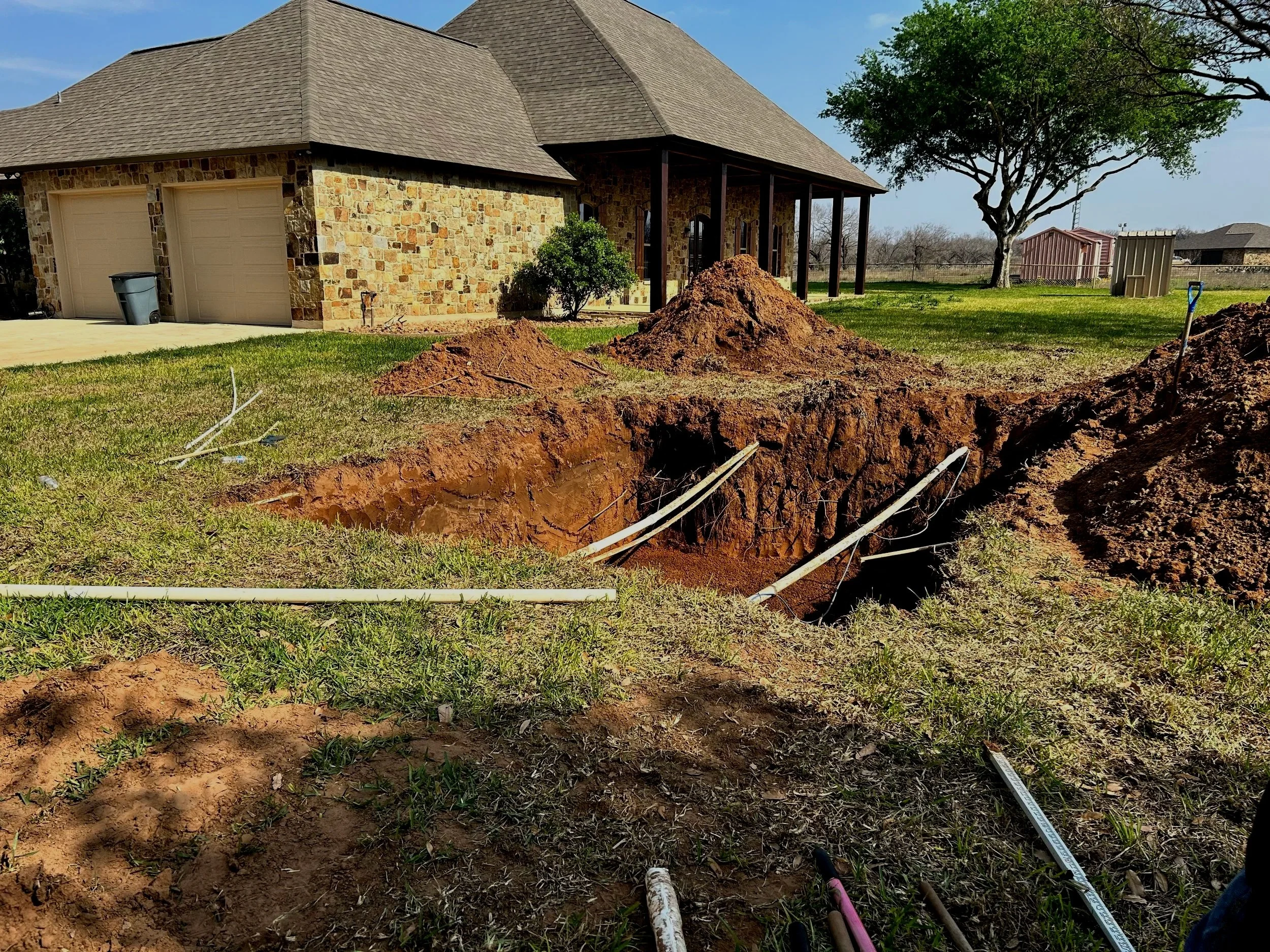 Construction site in the backyard of a house shows a large rectangular hole in the ground with pipes and wires. The house has a stone exterior, a sloped roof, and an attached covered porch. There is a pile of dirt near the hole, and a shovel is leani
