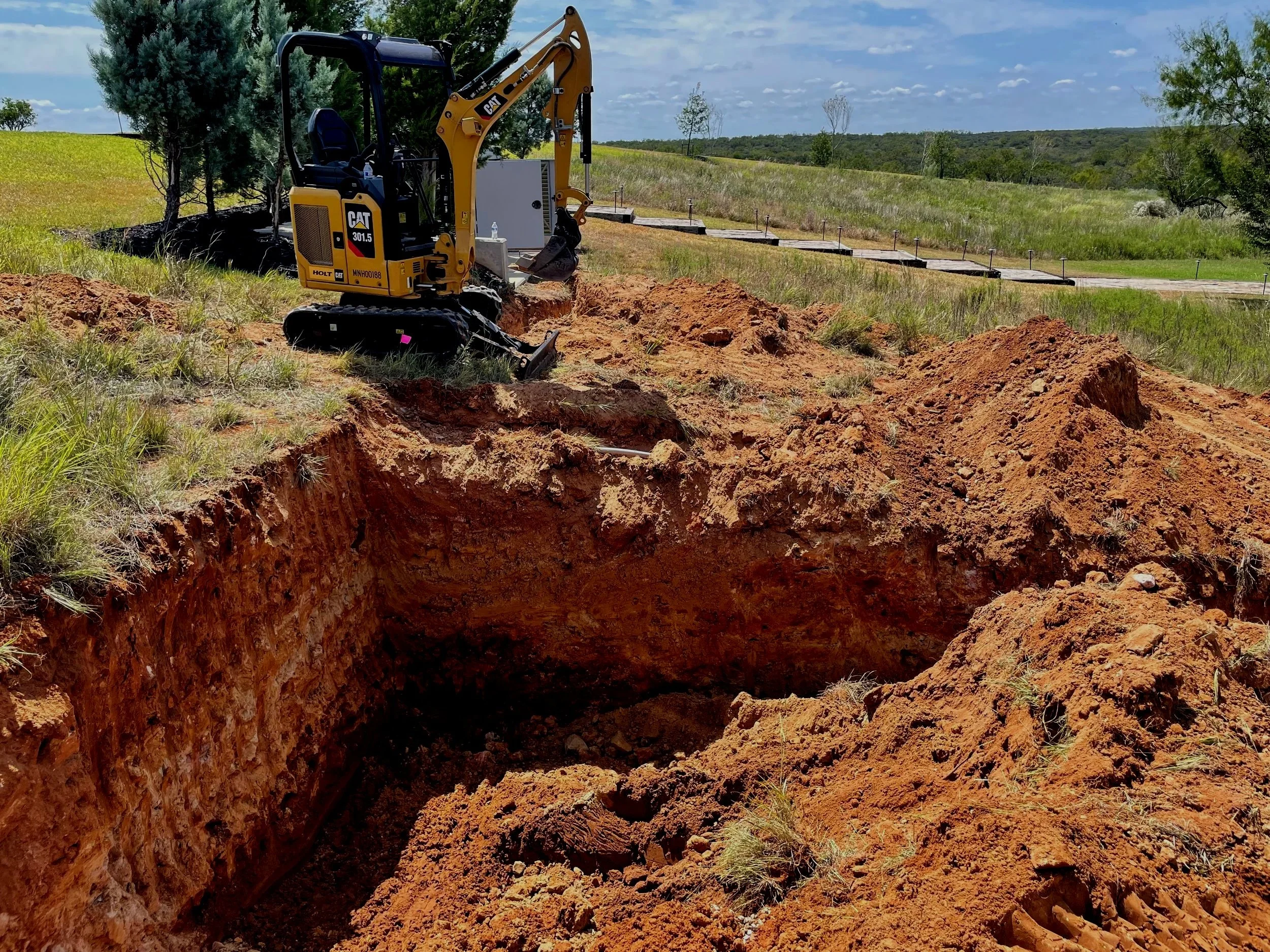 Construction site with a small yellow Caterpillar excavator digging a large hole, surrounded by grassy fields and trees under a partly cloudy sky.