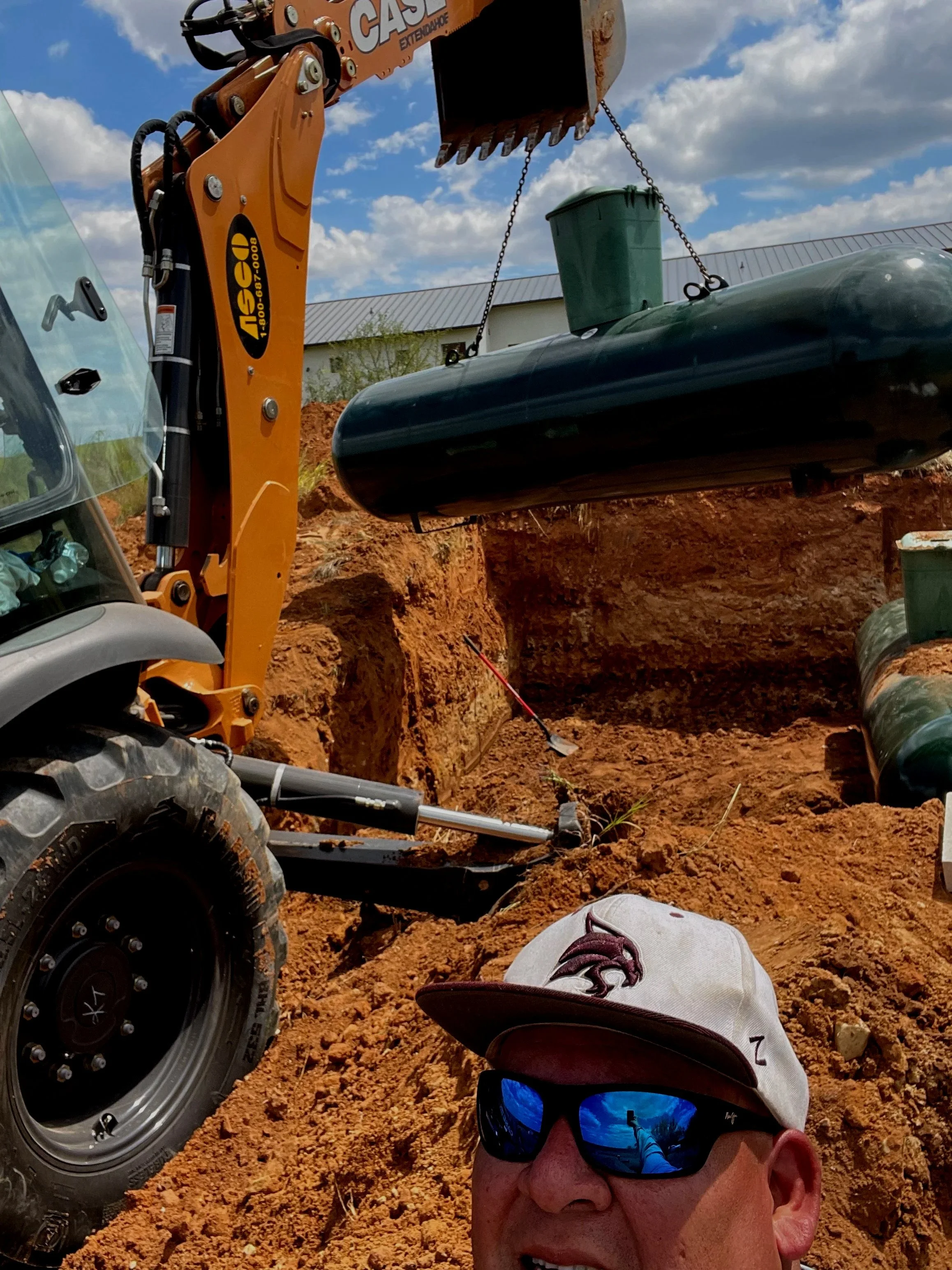 Person wearing a hat and sunglasses taking a selfie at a construction site with an excavator lifting a green trash bin. The background shows a partly cloudy sky and a building.