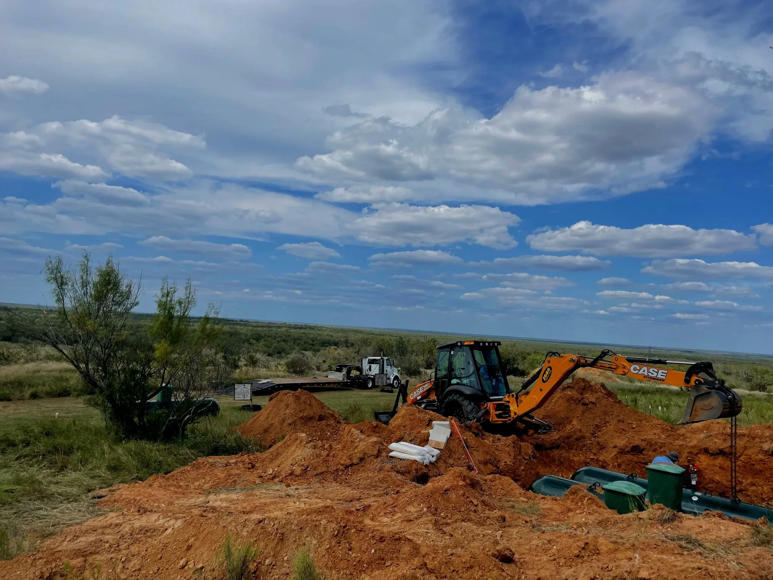 Construction site with a yellow CASE backhoe digging in red dirt, green vegetation, and blue sky with clouds, and a wide open landscape in the background.