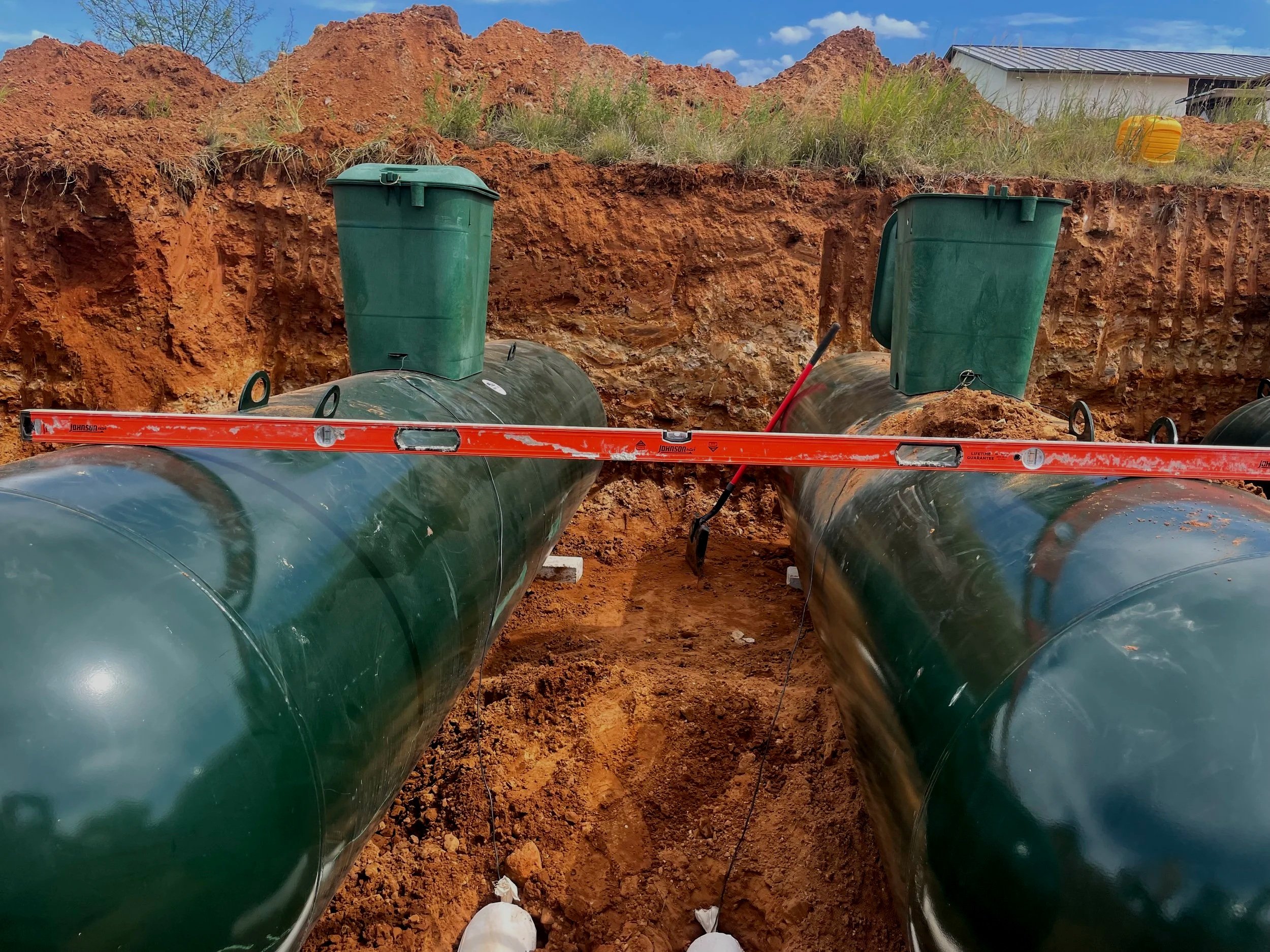 Underground installation of large green plastic pipes with soil and grass above, construction level and tools nearby, and a building in the background.