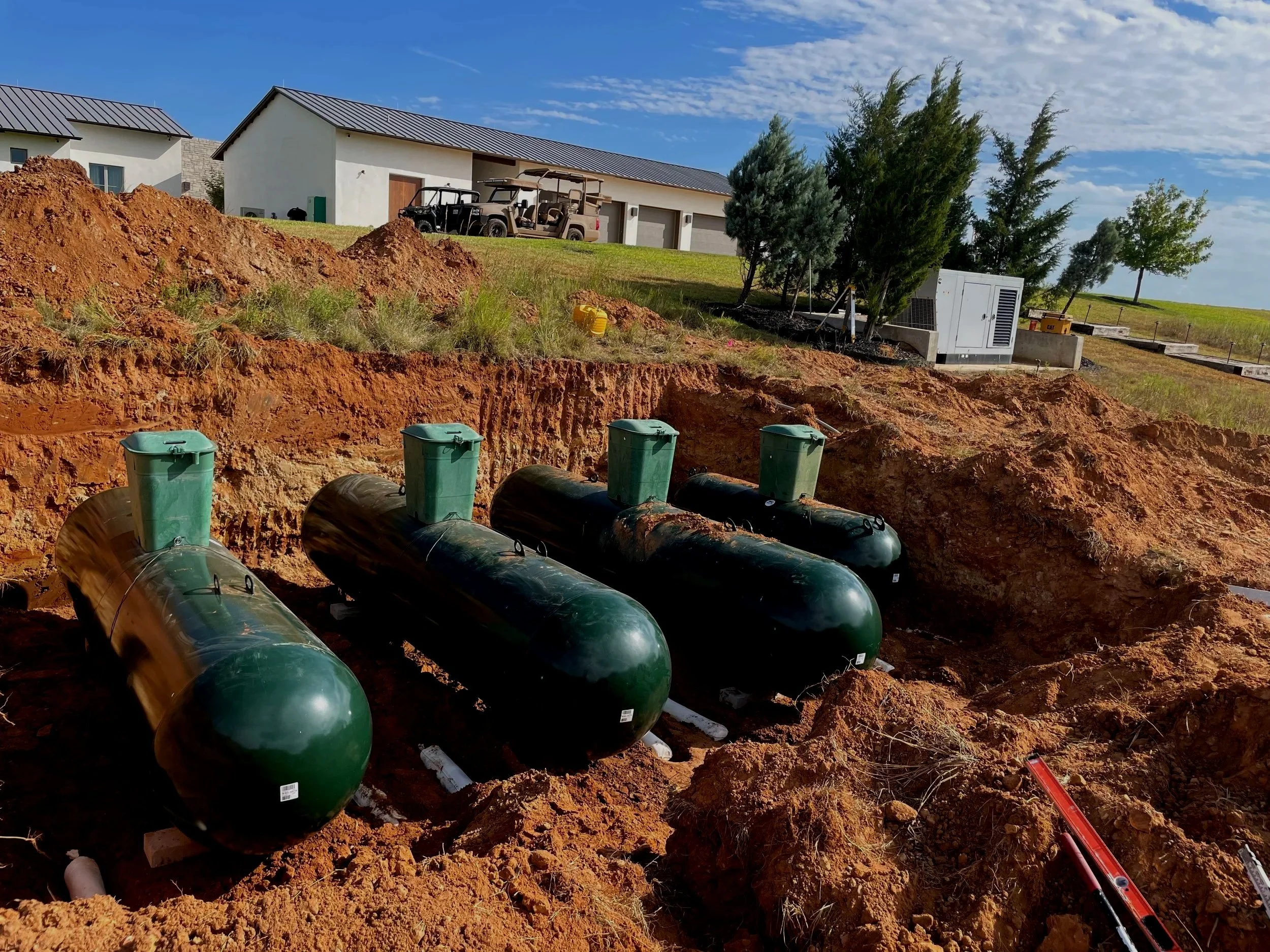Construction site with large underground pipes, dirt piles, and trees near modern houses under a clear blue sky.