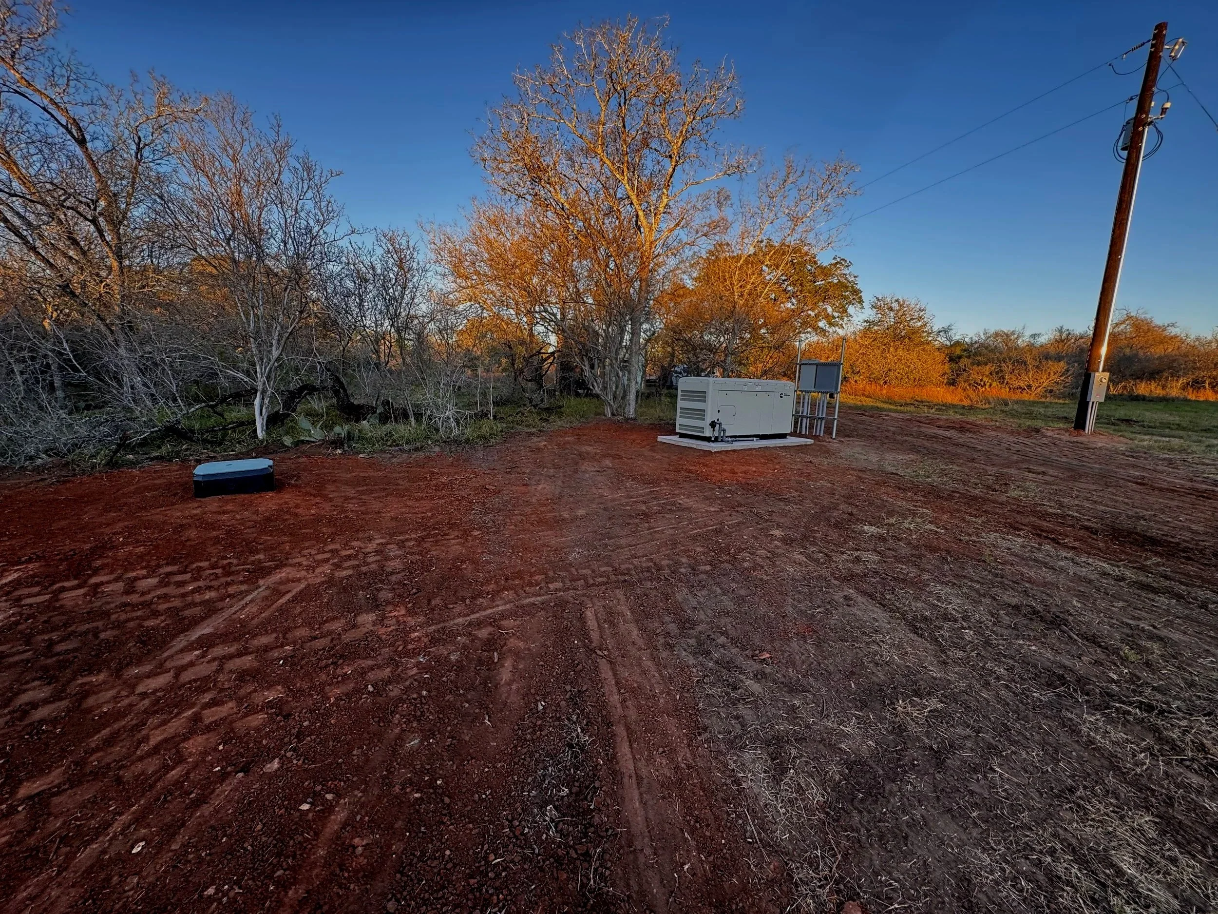 A dirt road with tire tracks, dry grass, and leafless trees in the background, alongside a utility pole and electrical equipment, under a clear blue sky at sunset.