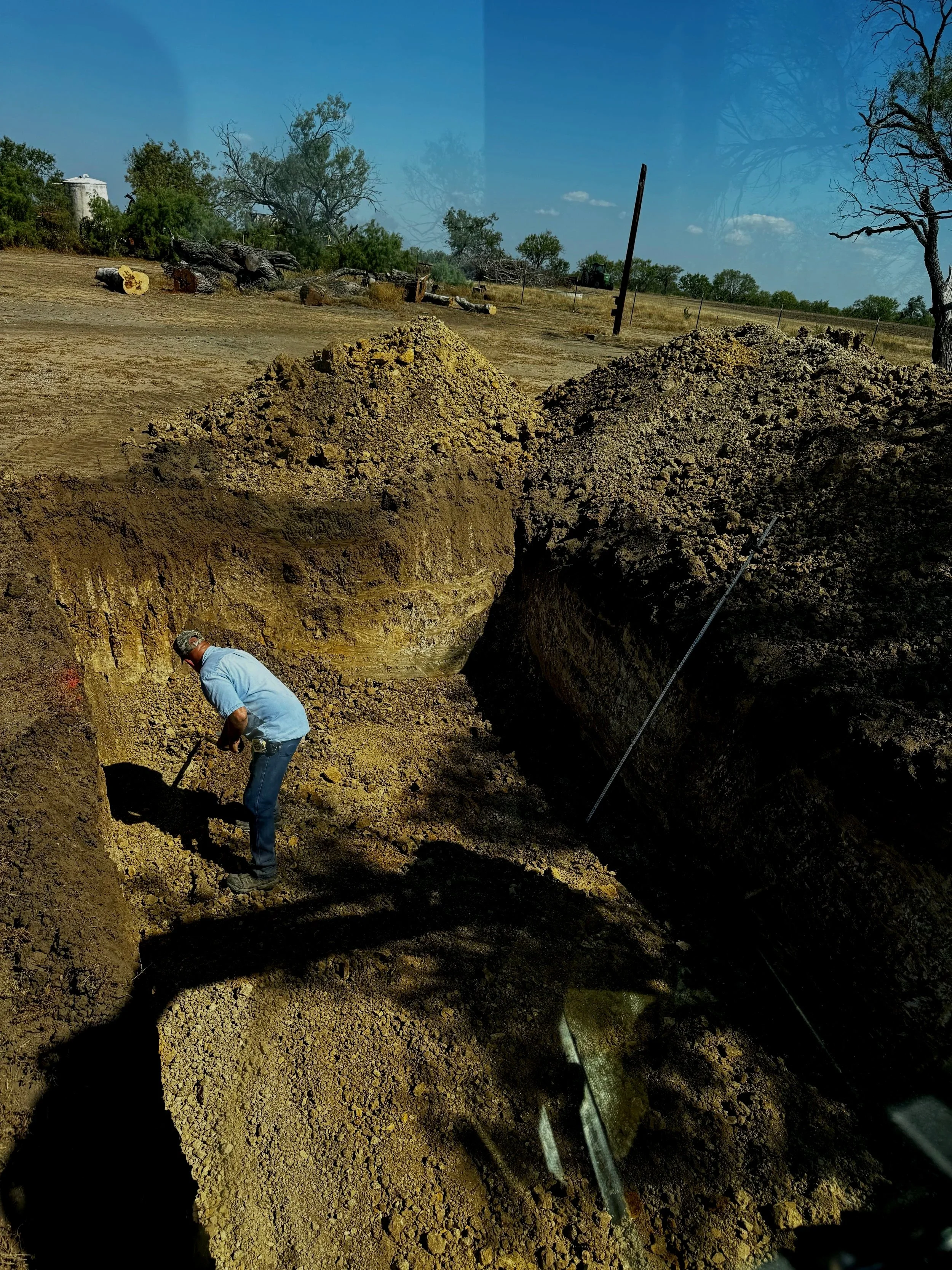 A man digging in a large excavation site with dirt piles and trees in the background.