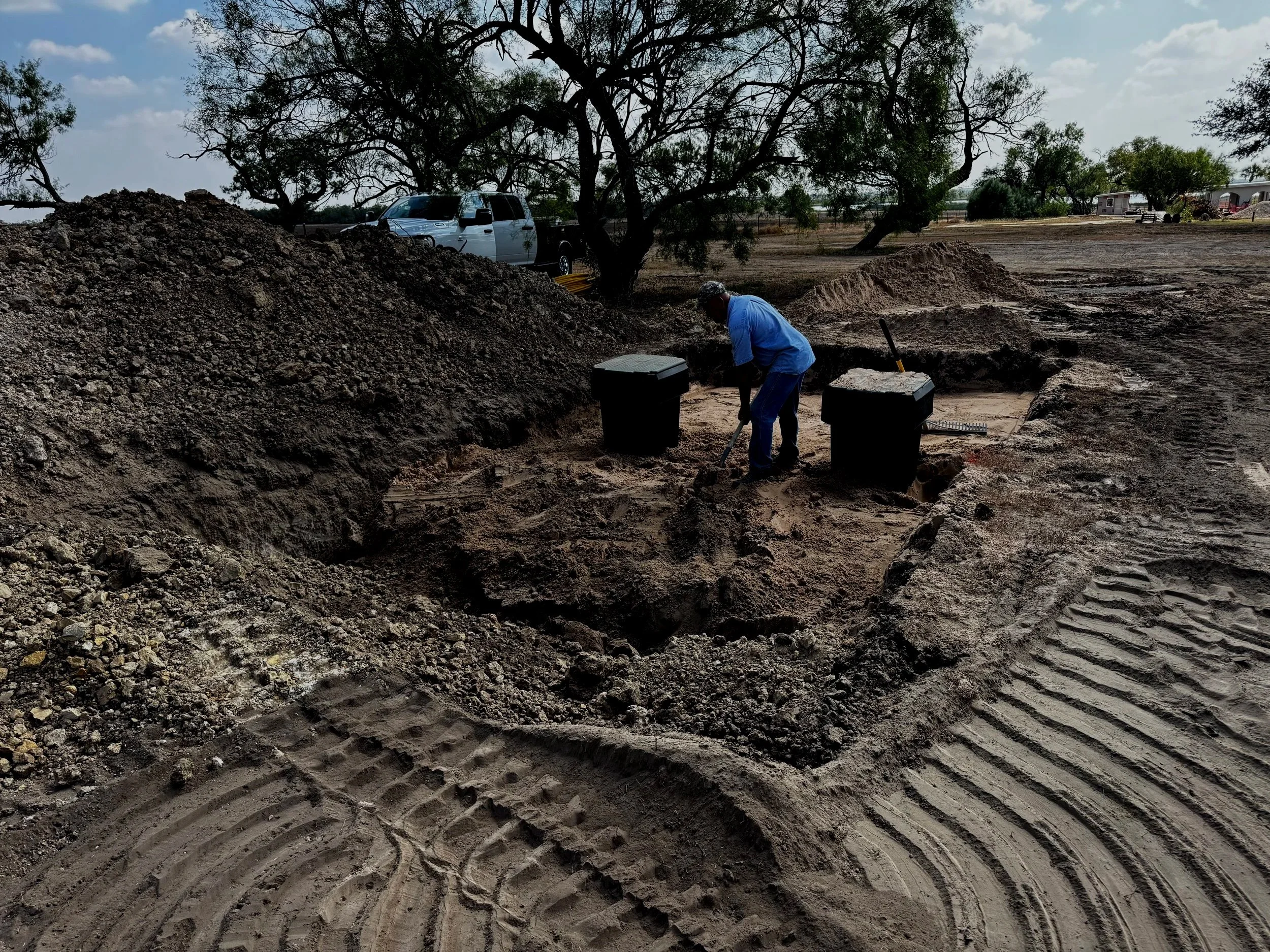 A person working in a dug-out area of dirt with large mounds of soil nearby. In the background, there is a white pickup truck parked under a tree, with a trees, a trailer, and some buildings visible further away.