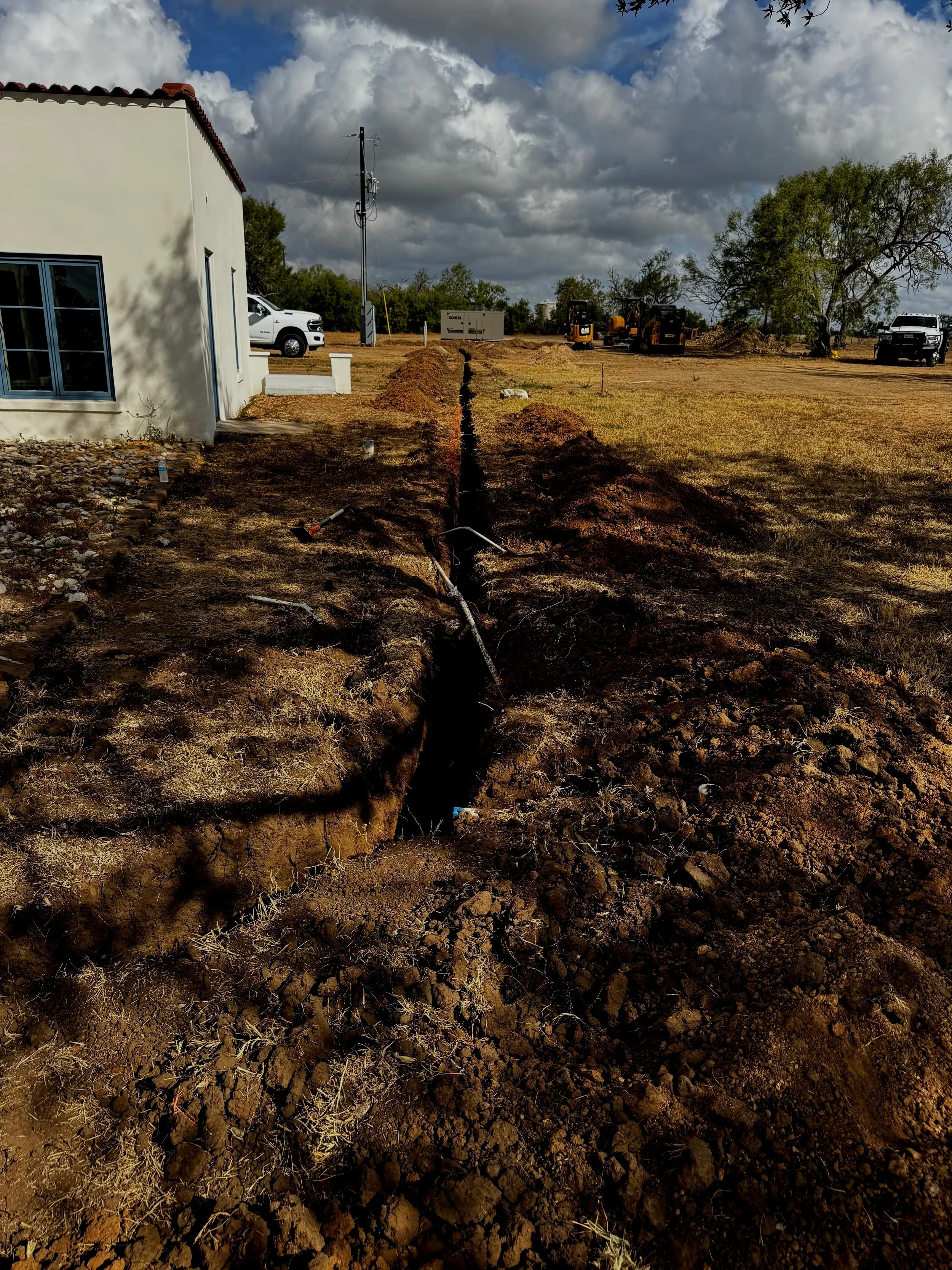A construction site with a trench dug in the ground, a white building on the left, construction equipment in the background, and a cloudy sky overhead.