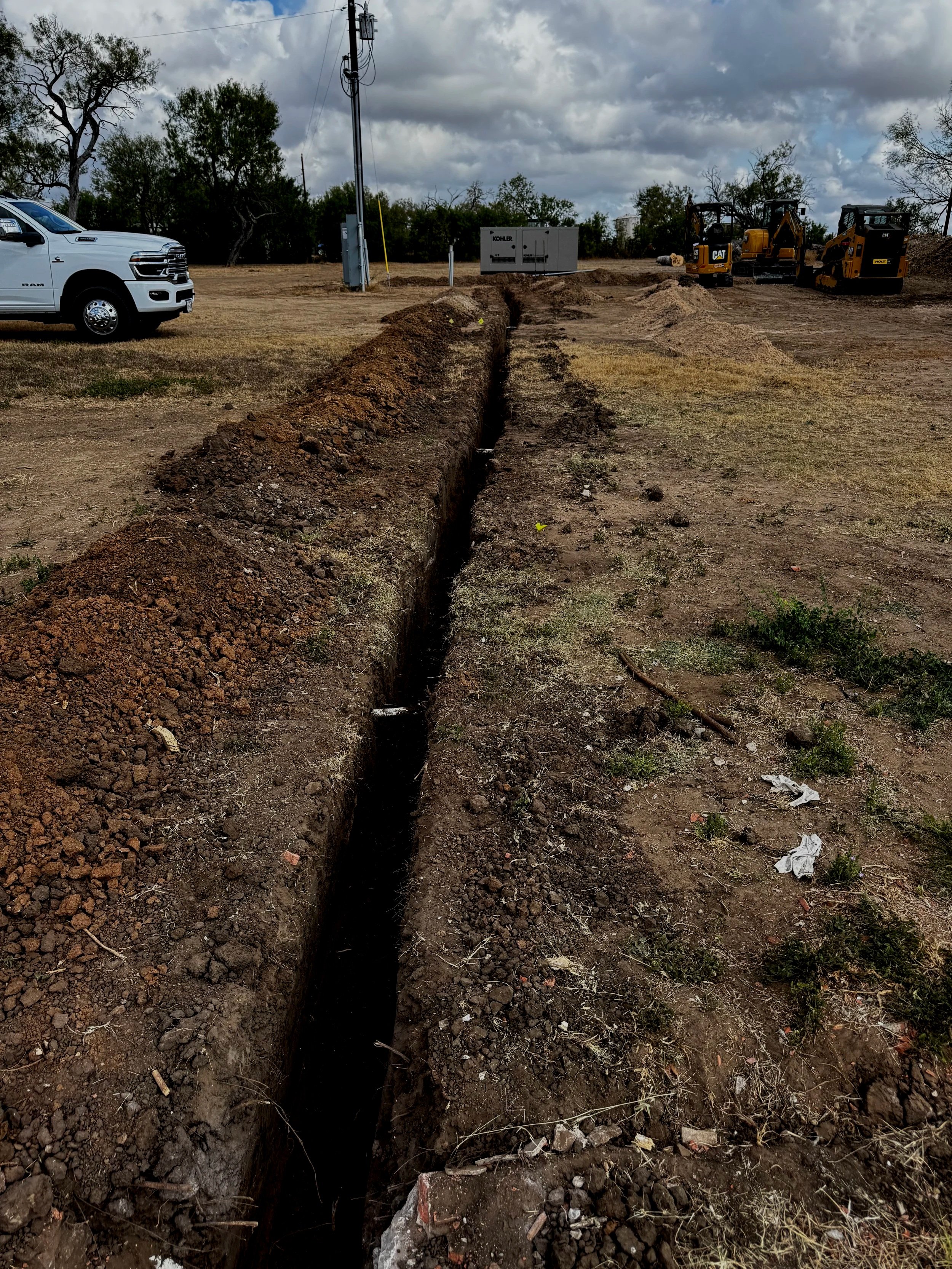 A construction site with a trench in the dirt, a white truck on the left, and small yellow excavators on the right. There are trees and cloudy skies in the background.
