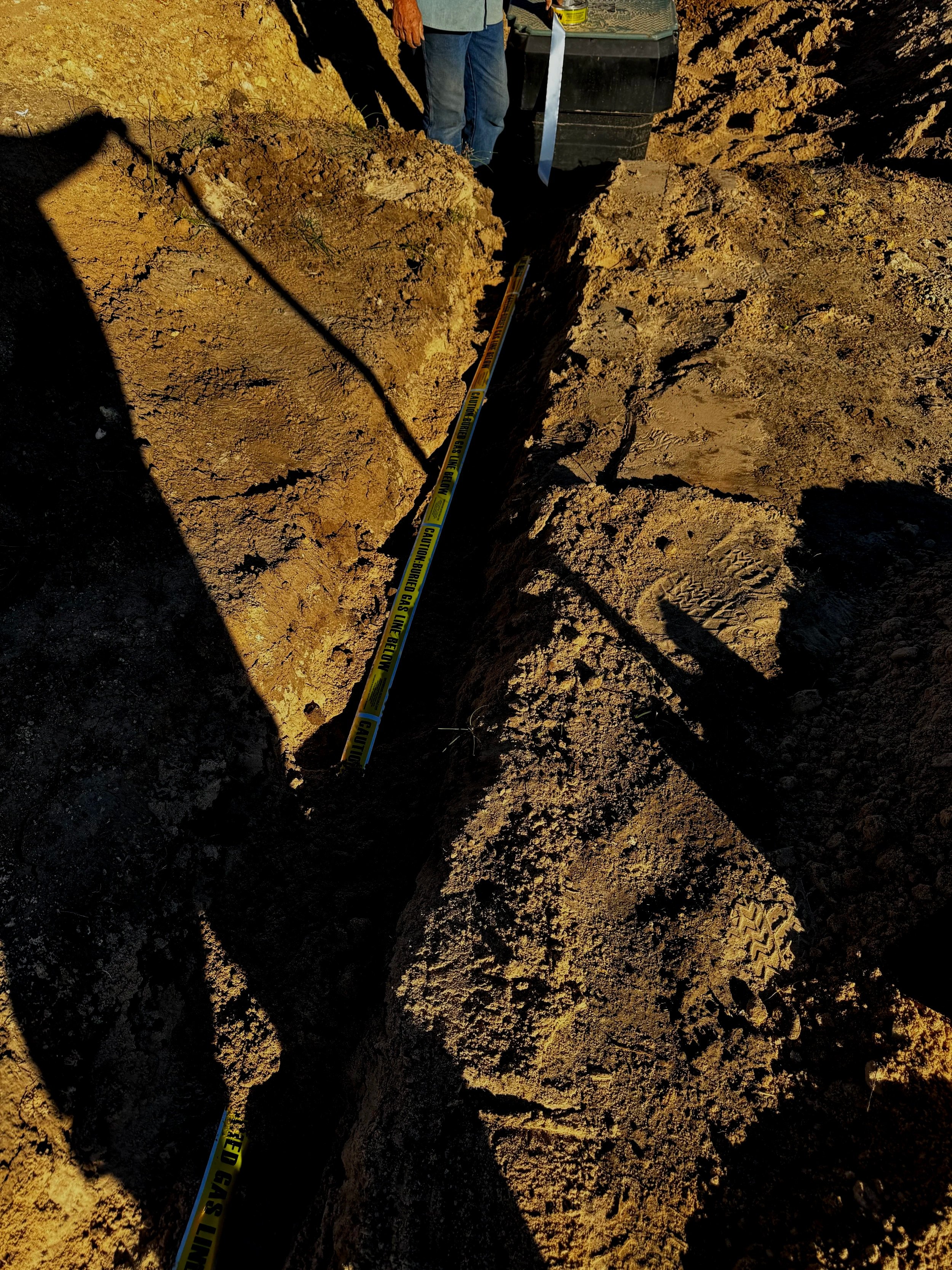A person standing in a deep excavation trench with dirt walls, with a yellow caution tape running along the trench.