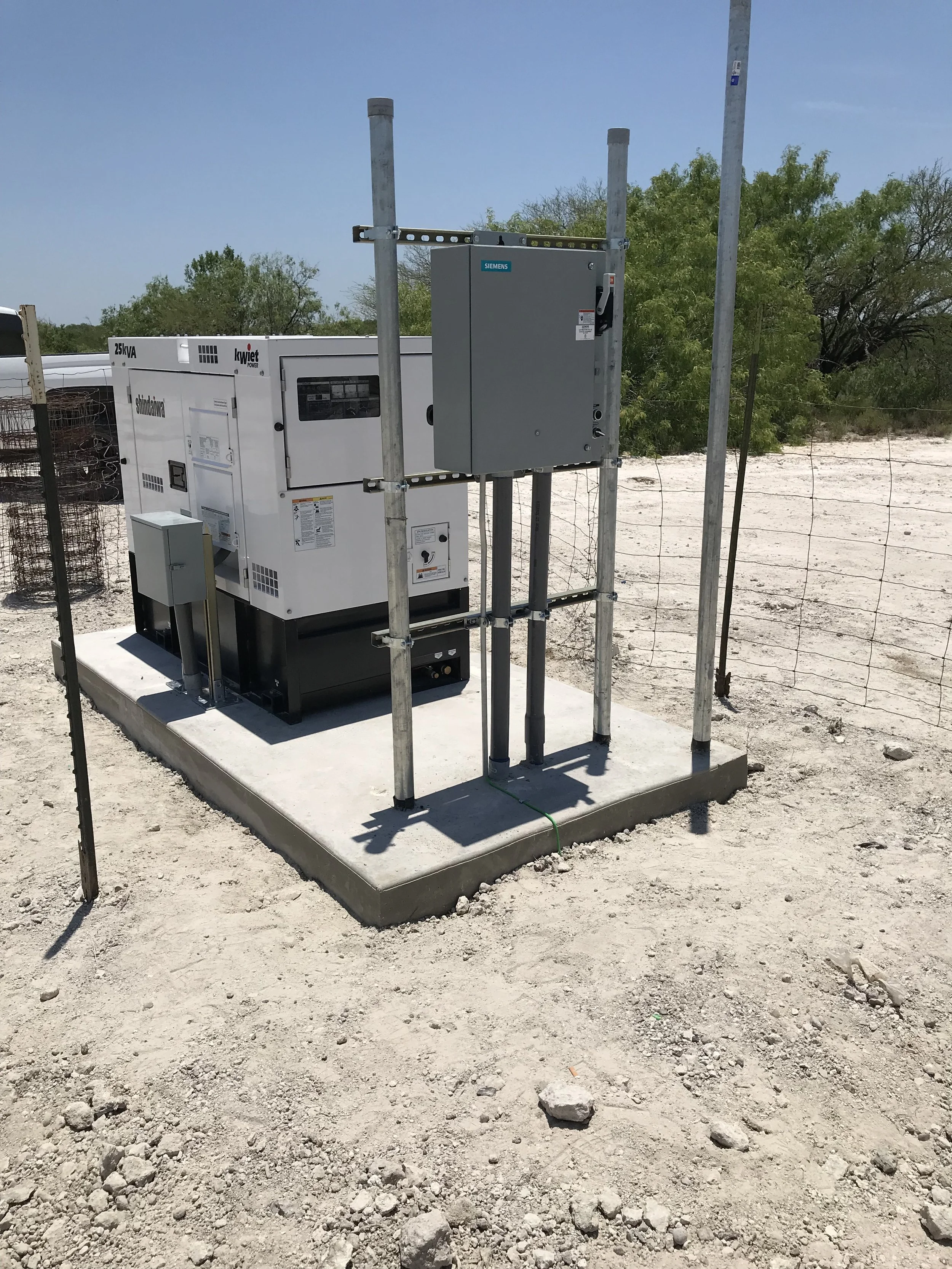 Outdoor electrical or utility electrical box installed on a concrete pad, surrounded by a metal safety fence, with trees and a clear blue sky in the background.