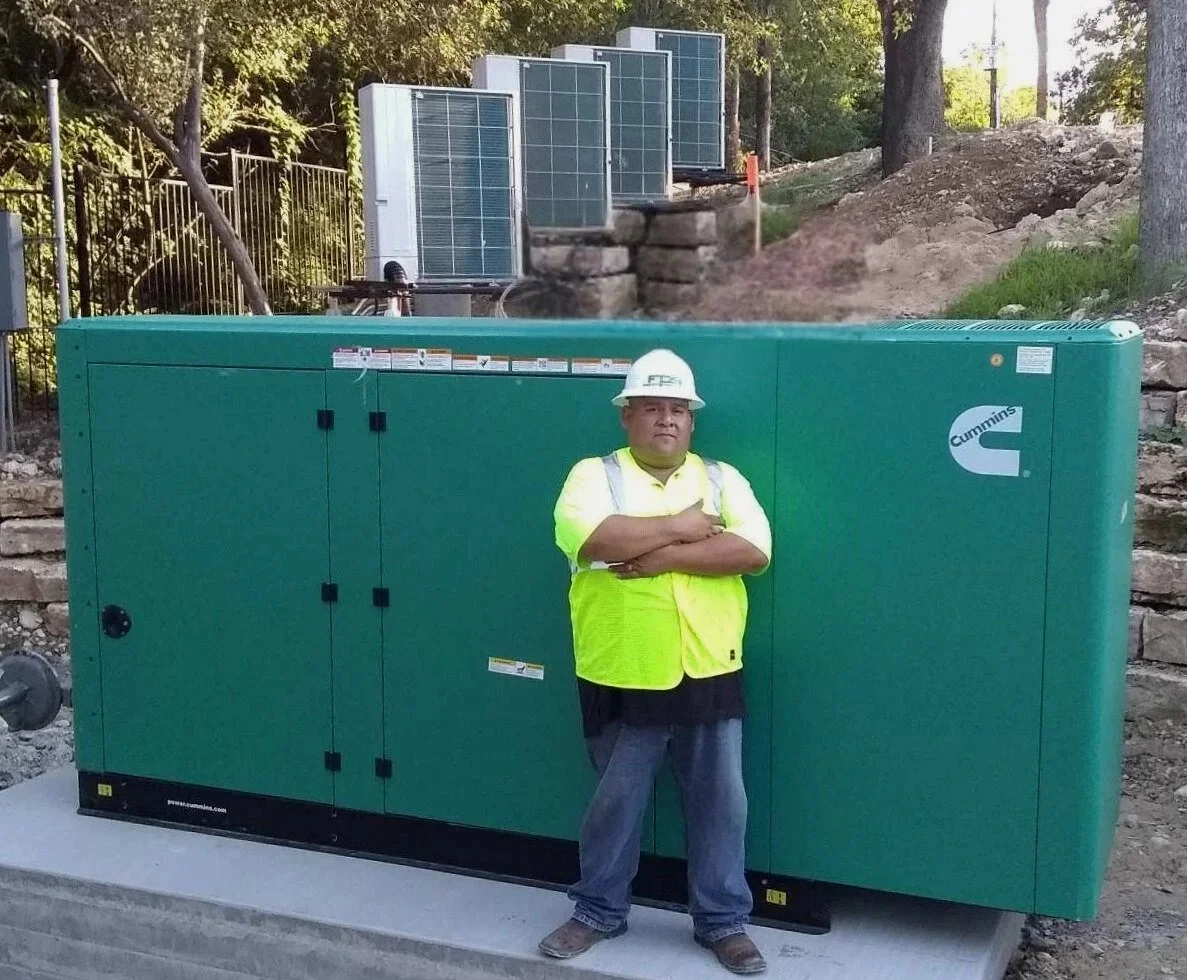 A man wearing a white hard hat and yellow safety vest standing with arms crossed in front of a large green generator with the brand name Cummins on it. Construction site outdoors with trees and equipment in the background.