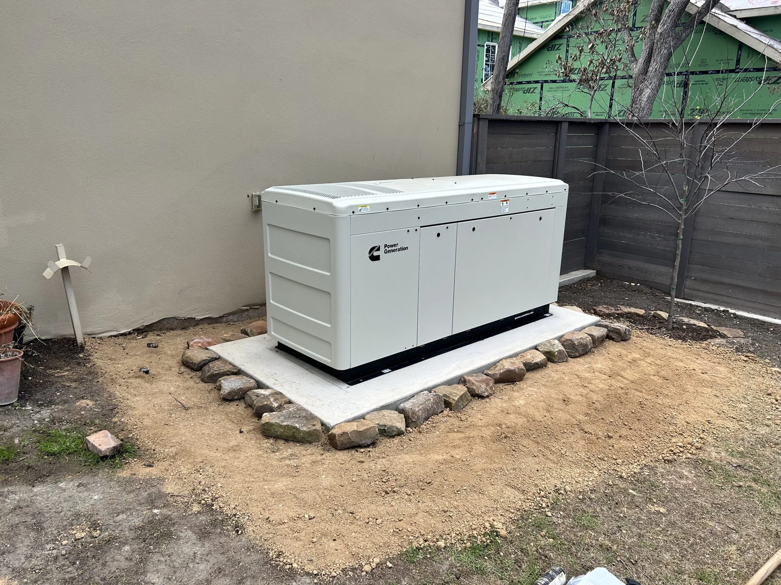 An outdoor scene showing a gray power generator on a concrete pad, with a small leafless tree to the right, surrounded by dirt and scattered rocks, with a beige stucco wall on the left and a wooden fence with construction in the background.