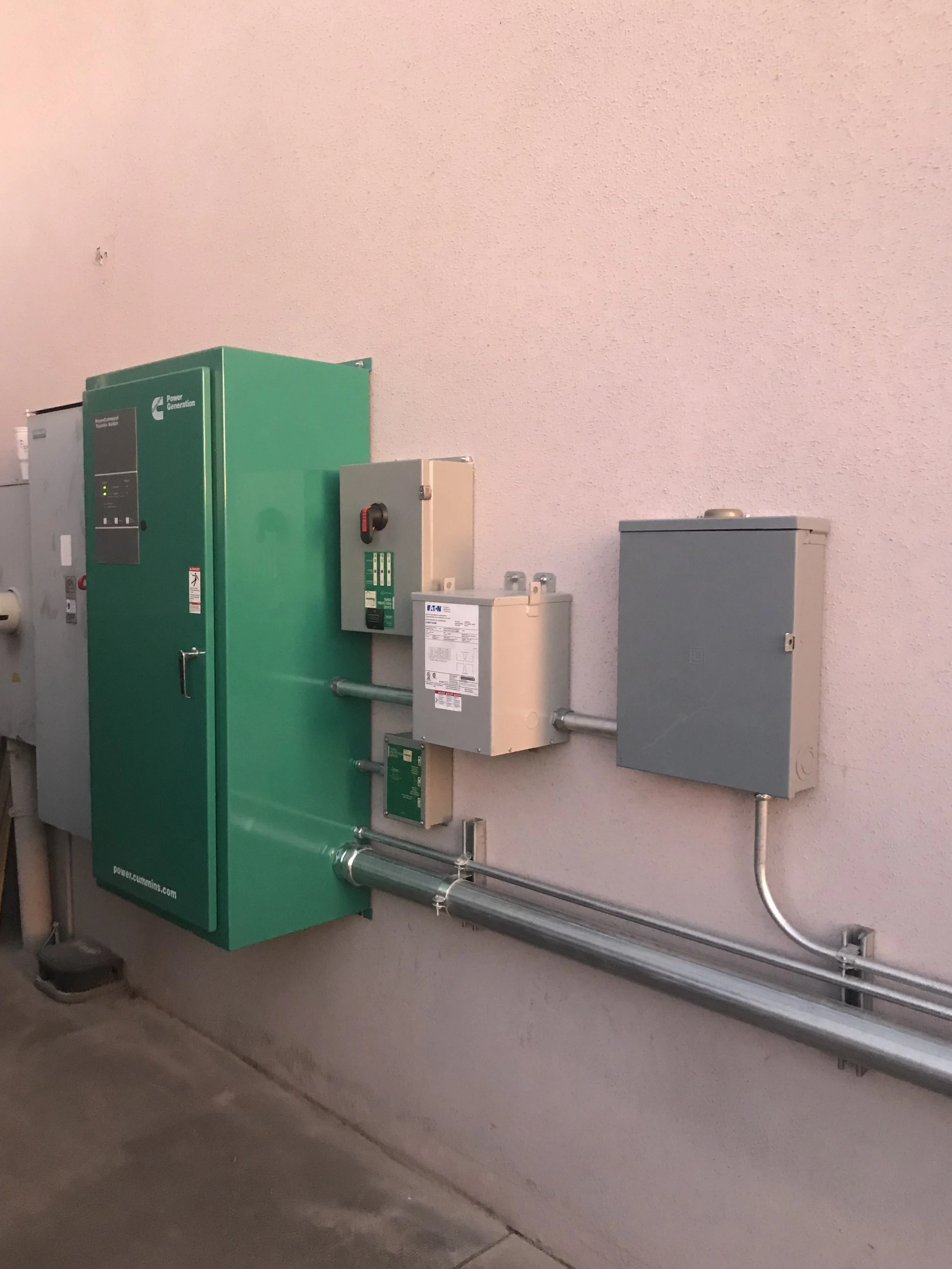Electrical and utility boxes mounted on a pink-textured wall with metal conduit piping beneath.