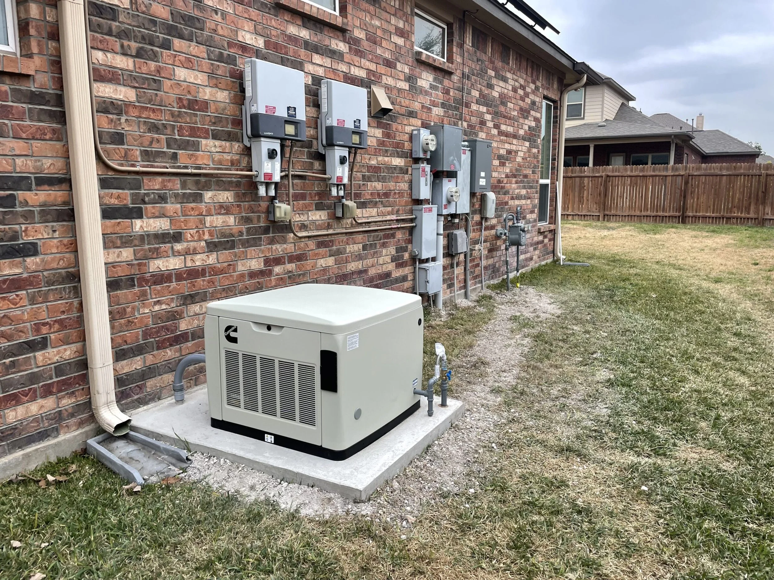 Outside view of a house brick wall with electrical meter boxes, a generator, and utility connections, next to a grassy yard and wooden fence under cloudy sky.