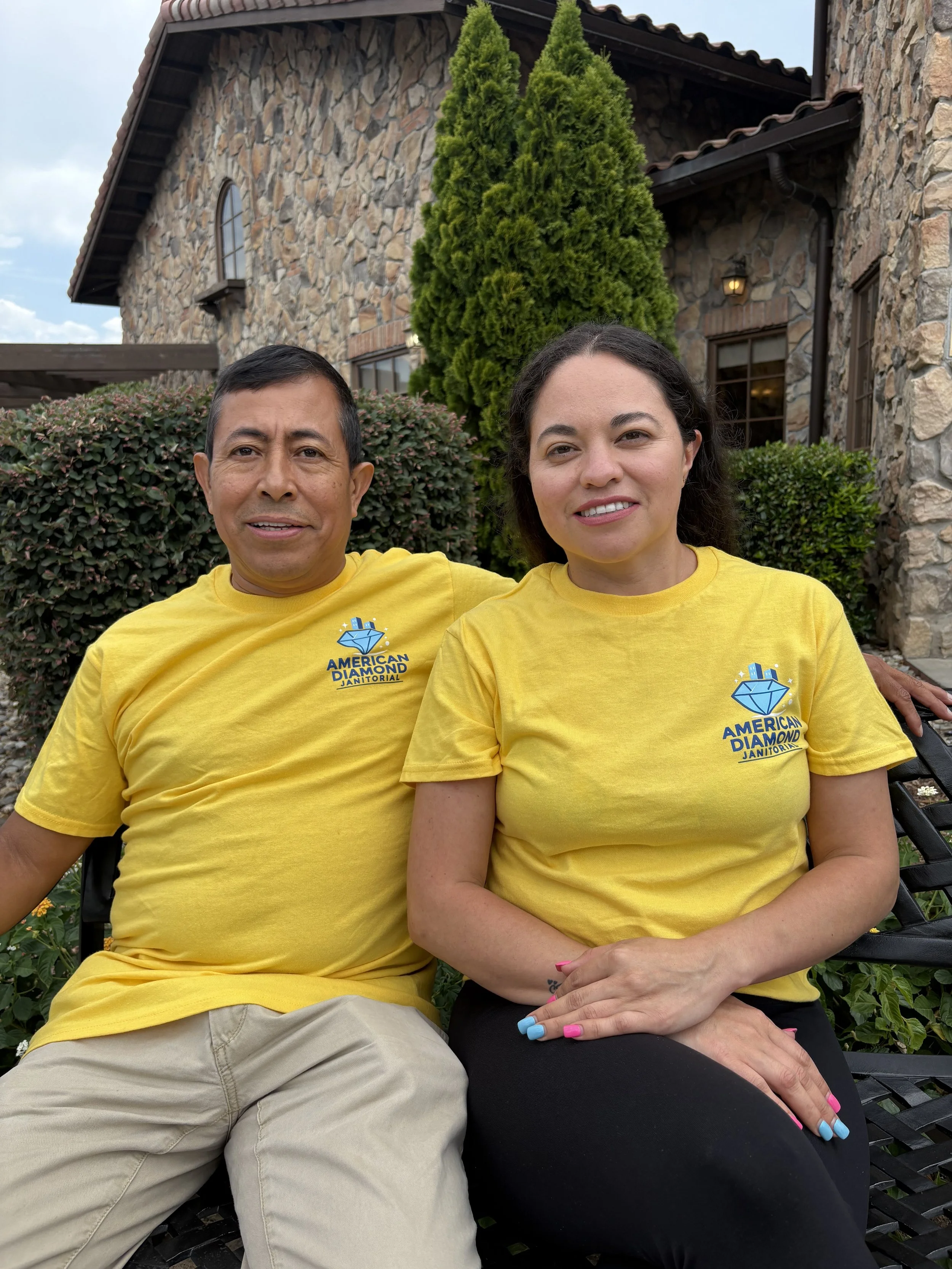 A man and woman sitting on a bench outdoors in front of a stone house, both wearing bright yellow T-shirts with a blue diamond logo and the text 'American Diamond Janitorial.' The man has short black hair, and the woman has long dark hair and colorful nails.