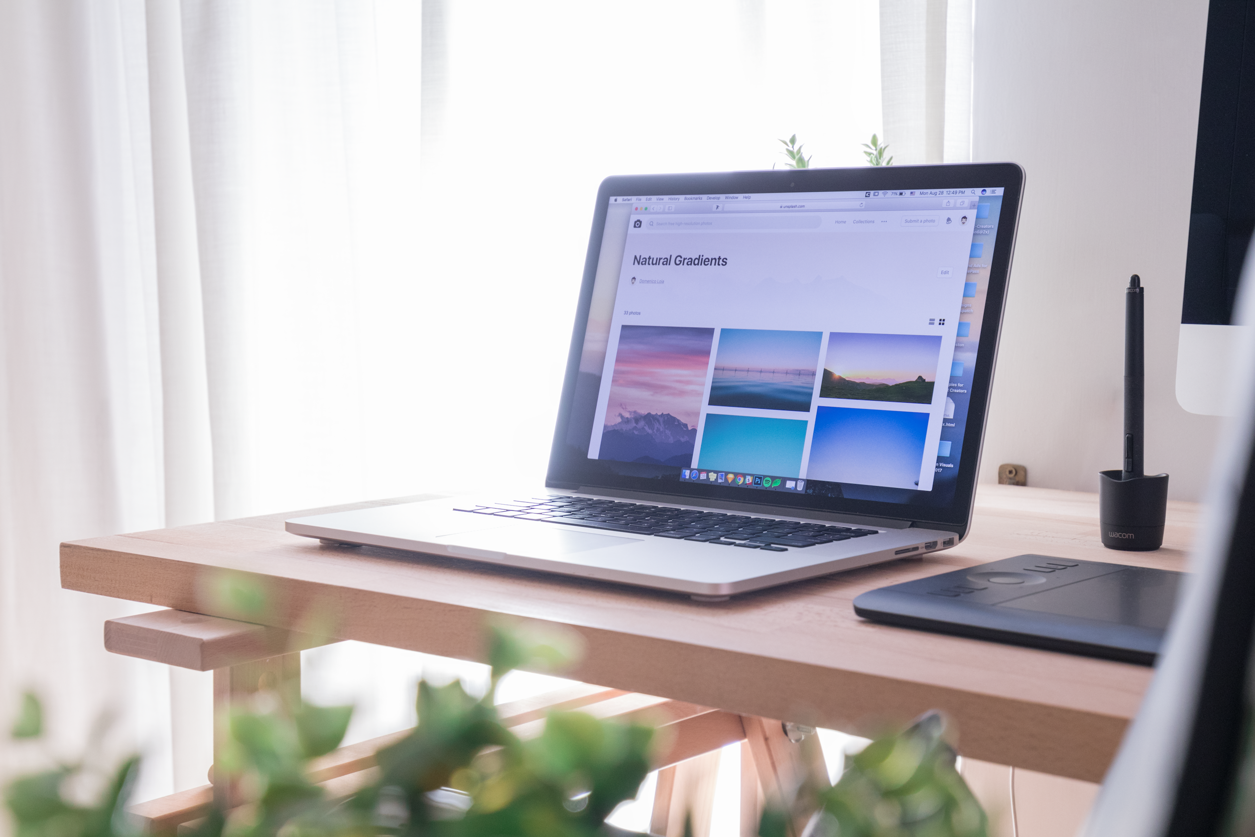 A laptop on a wooden desk displaying a photo management website with colorful gradient images. A graphics tablet and a pen stand are also on the desk, with sunlight coming through sheer curtains in the background.
