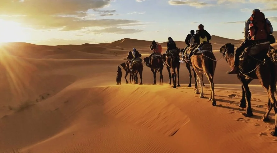 Group of people riding camels through a desert during sunset, with sand dunes and a partly cloudy sky in the background.