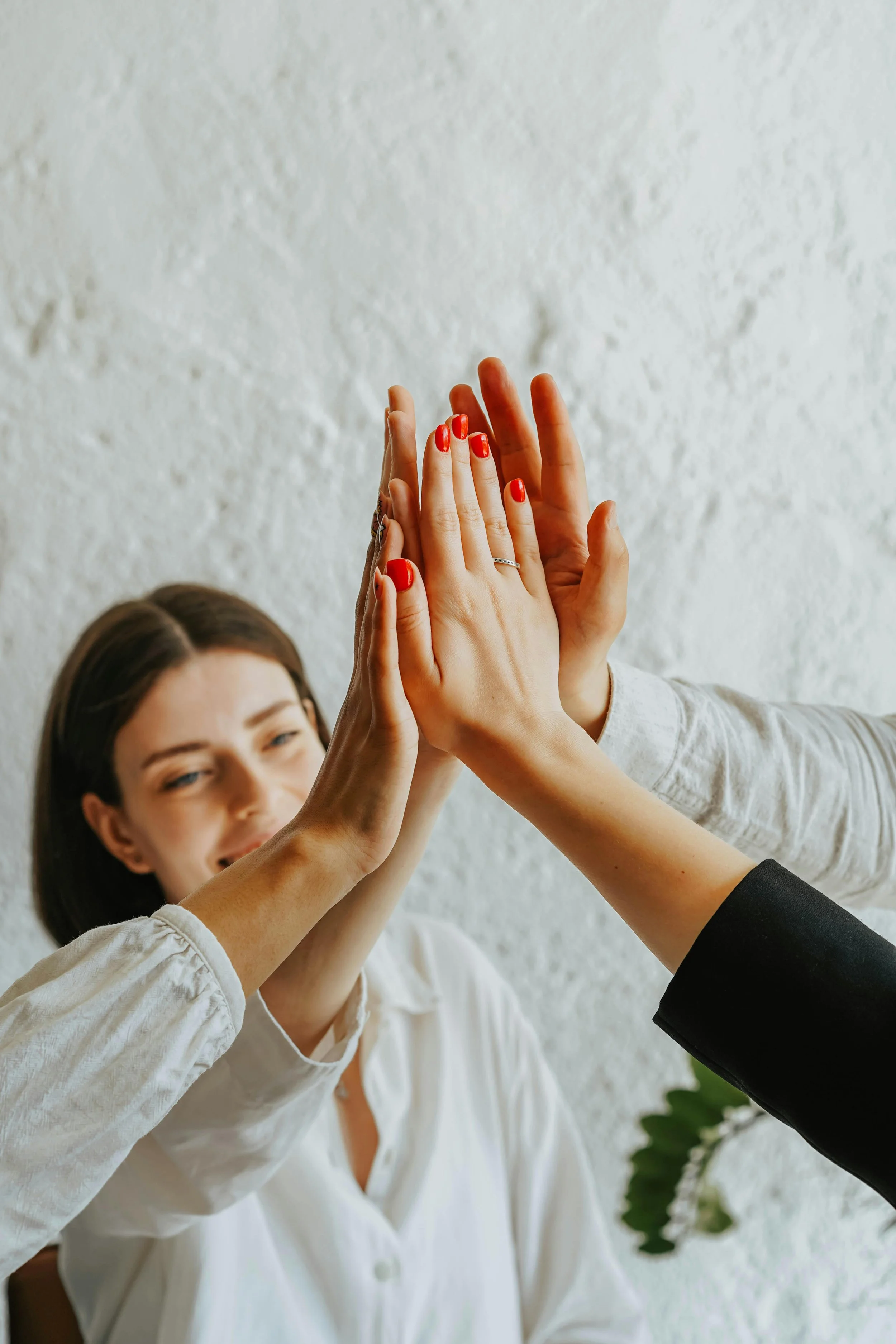 Three women giving a high five together, celebrating, with a textured white wall in the background.