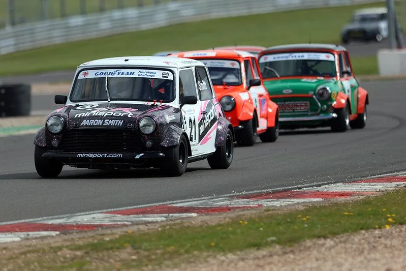 A vintage blue racing car with the number 89 on the side, driving on a racetrack, with other cars visible in the background.