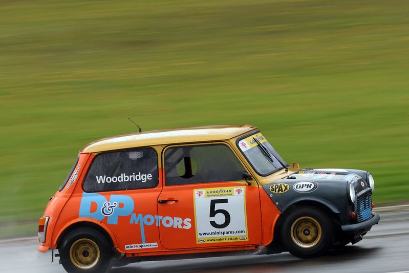 Vintage race car with the number 77 on the side, racing on a track, painted in red, white, and black with sponsorship decals.