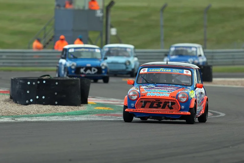 Three vintage Mini Cooper race cars on a racetrack, with two blue ones and a green one in the background.