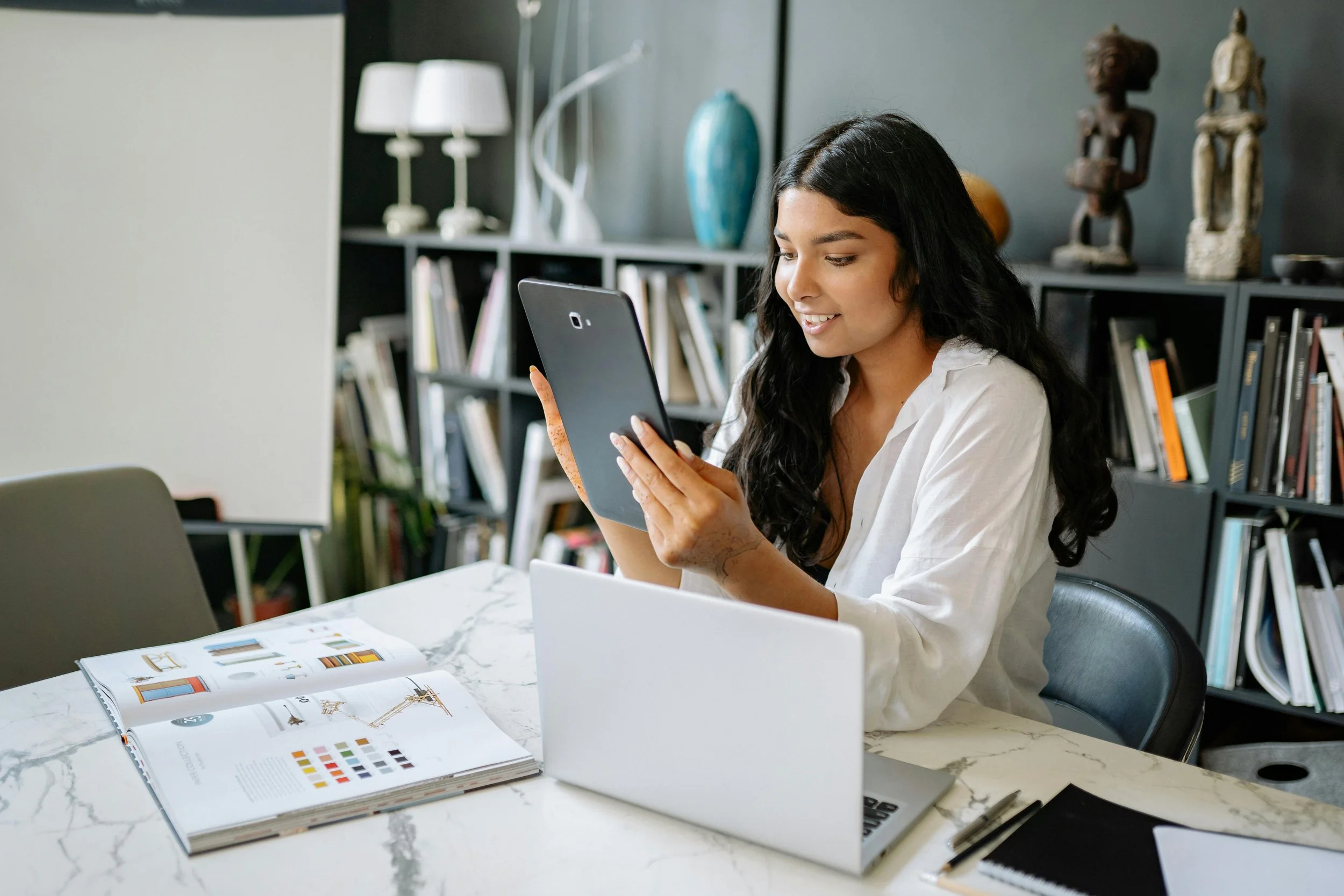 Woman sitting at a marble table, holding a tablet, with open magazine, laptop, and notebook with pens, in a room with bookshelves and decorative sculptures.