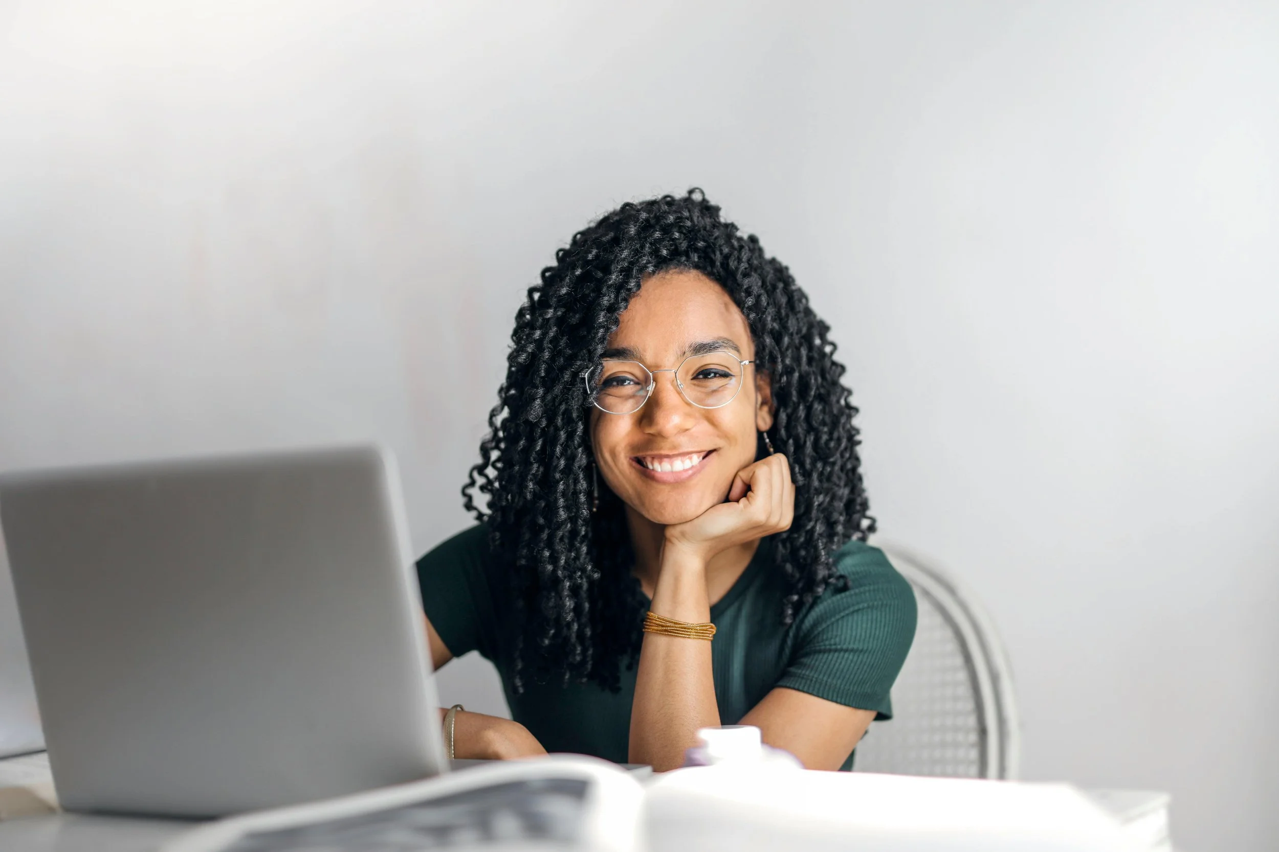 A woman with curly black hair wearing glasses, smiling, sitting at a desk with a laptop and papers, resting her chin on her hand.