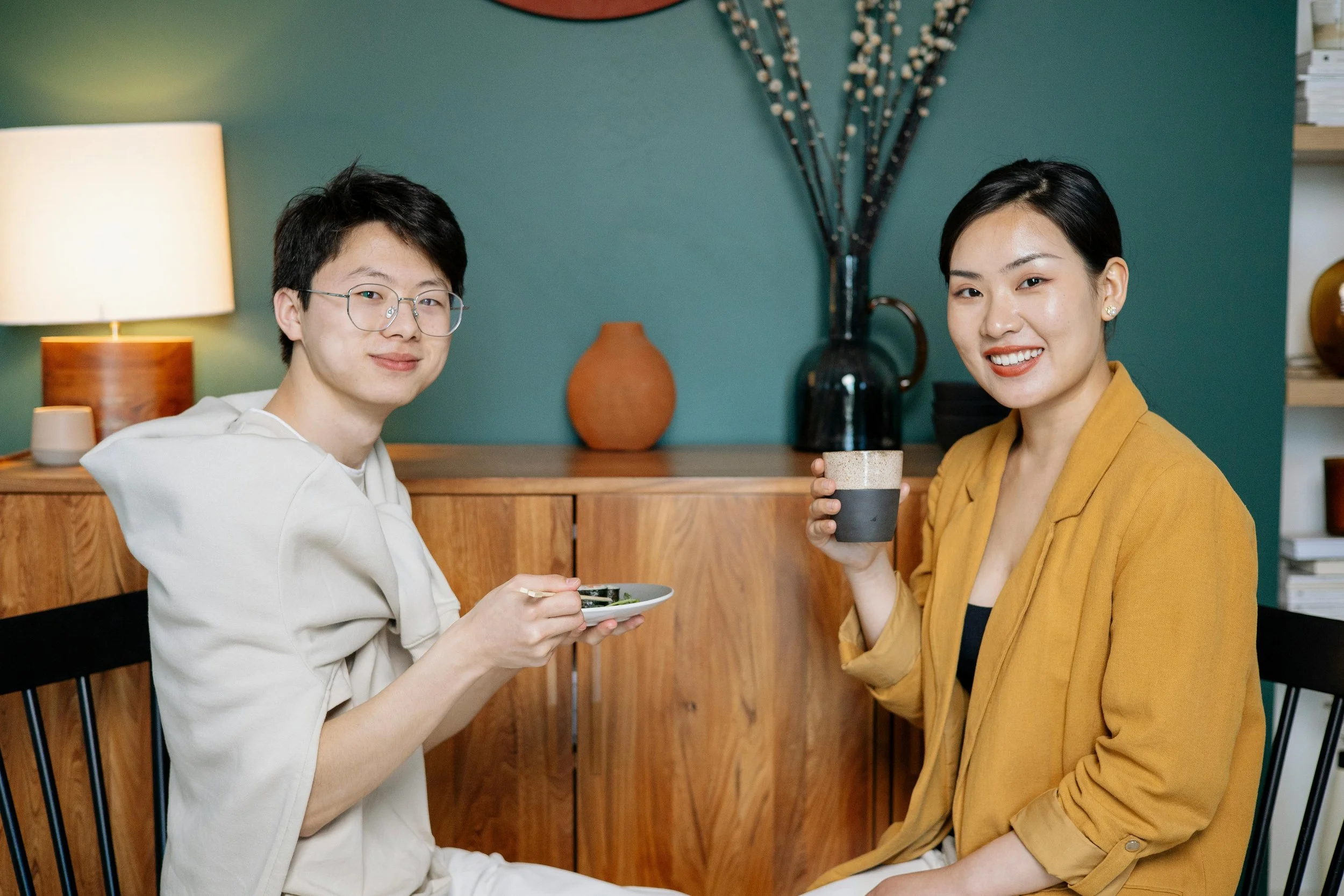 Two women sitting indoors, one holding a cup and the other with a small plate, smiling at the camera, with a wooden cabinet, vases, and a teal wall in the background.