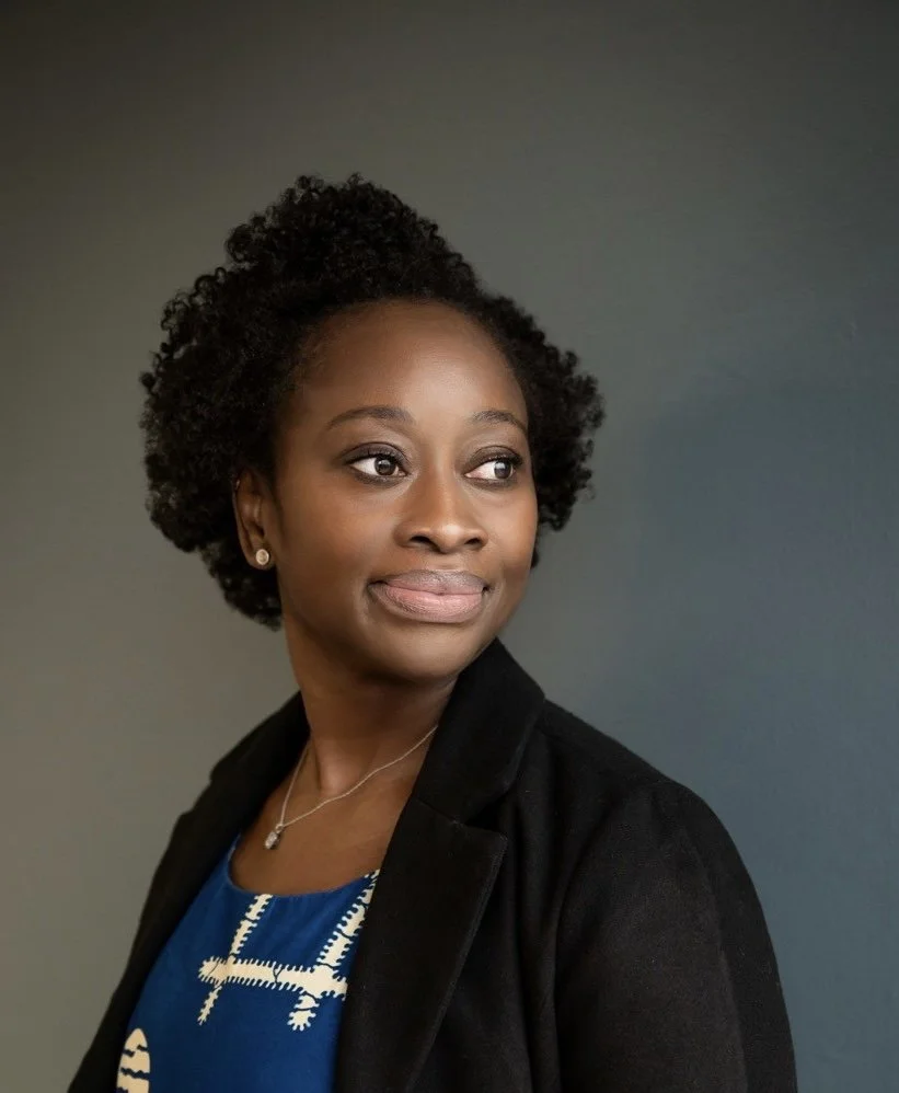 Portrait of a Black woman with curly hair, wearing earrings, a necklace, a blue top, and a black blazer, against a dark background.