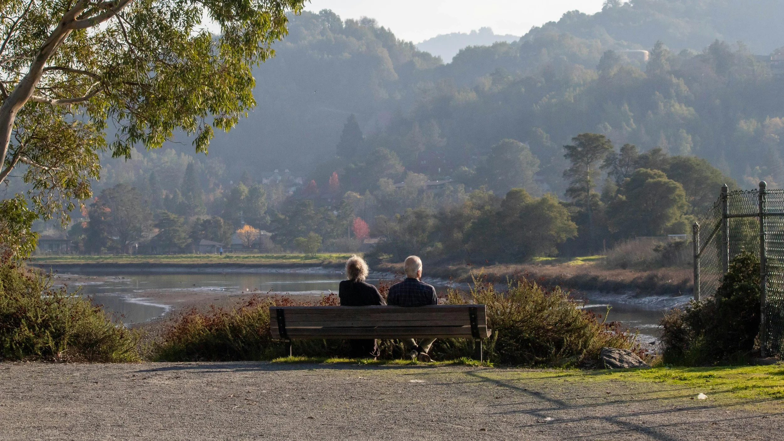 An elderly couple sits on a park bench near a river, facing away from the camera with mountains and trees in the background.