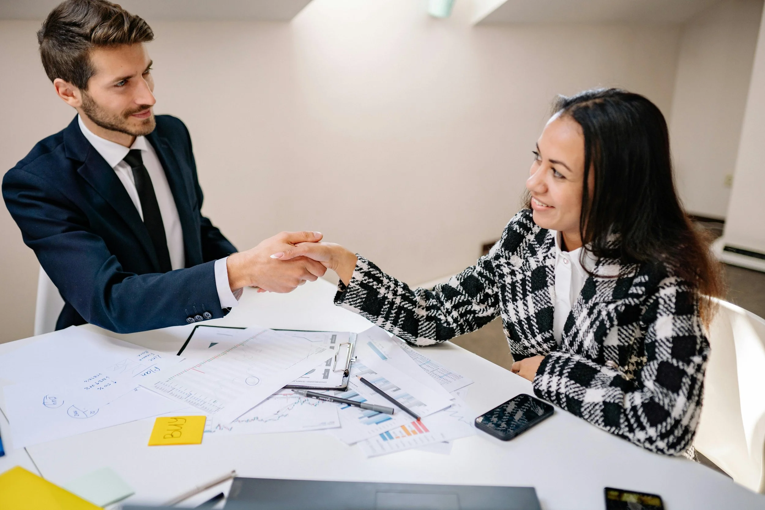 A man in a suit shaking hands with a woman in business attire at a table covered with documents and a smartphone.