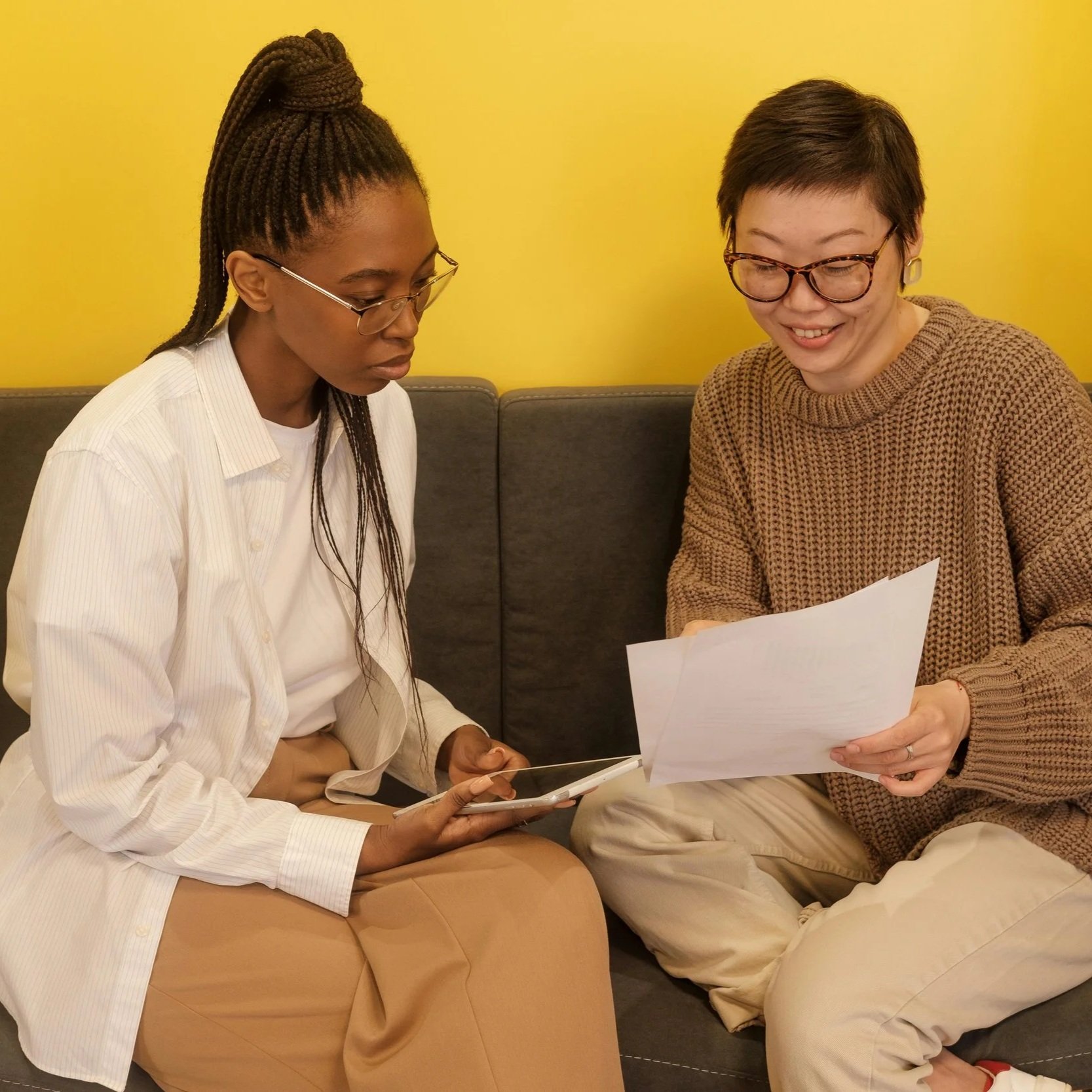 Two women sitting on a dark gray sofa with a yellow wall, looking at a paper and a tablet.