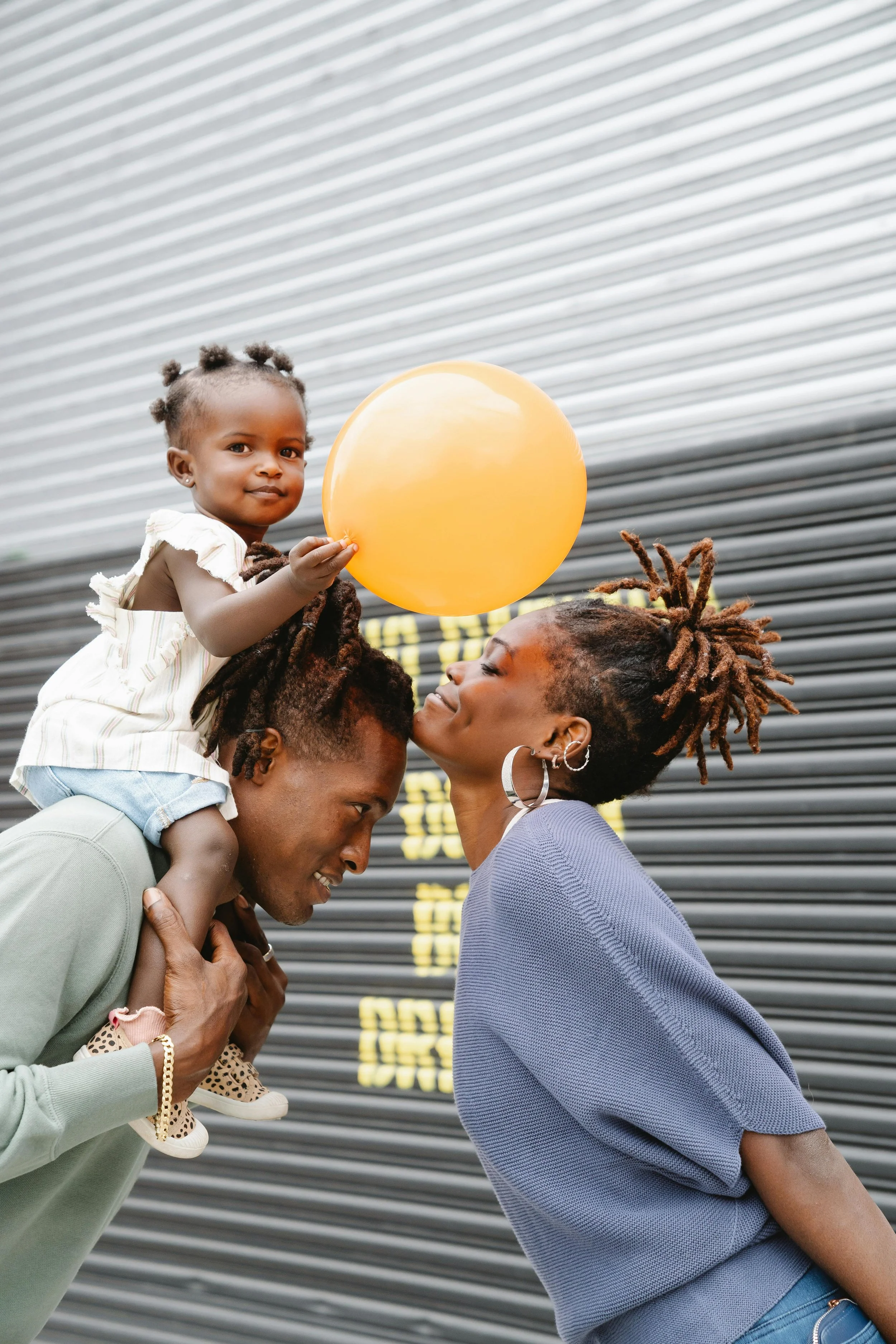 A family of three, consisting of a woman, a man, and a young girl, is enjoying time outdoors. The woman, with styled dreadlocks, large hoop earrings, and a blue top, is leaning close to the man. The man, with short hair and wearing a green long-sleeved shirt, is carrying the young girl on his shoulders. The girl is holding a yellow balloon and has her hair styled in small twists. They are standing in front of a dark, metallic rolling door with yellow text.