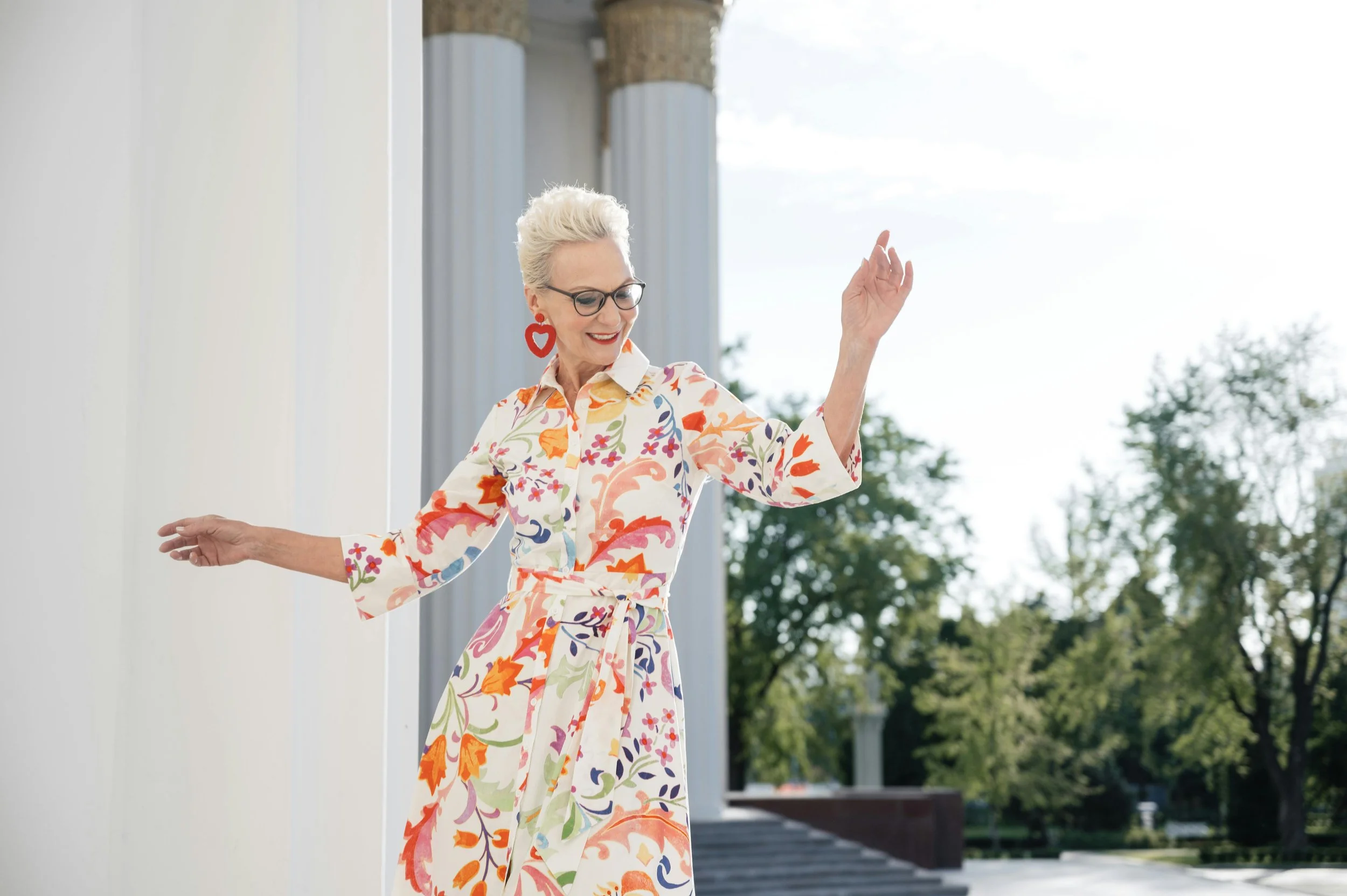 A woman with short, platinum blonde hair and glasses, wearing a colorful, floral dress with a belt, is smiling and dancing outdoors near classical columns with trees in the background.