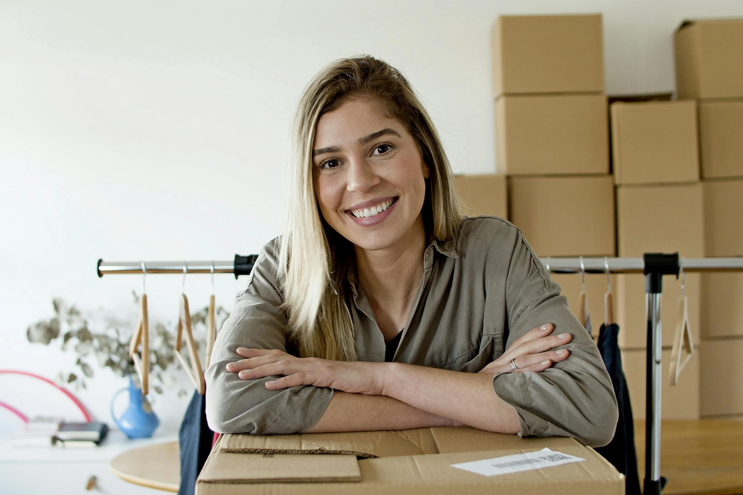 A woman smiling and sitting behind a cardboard box in a room with stacked boxes and a clothing rack.