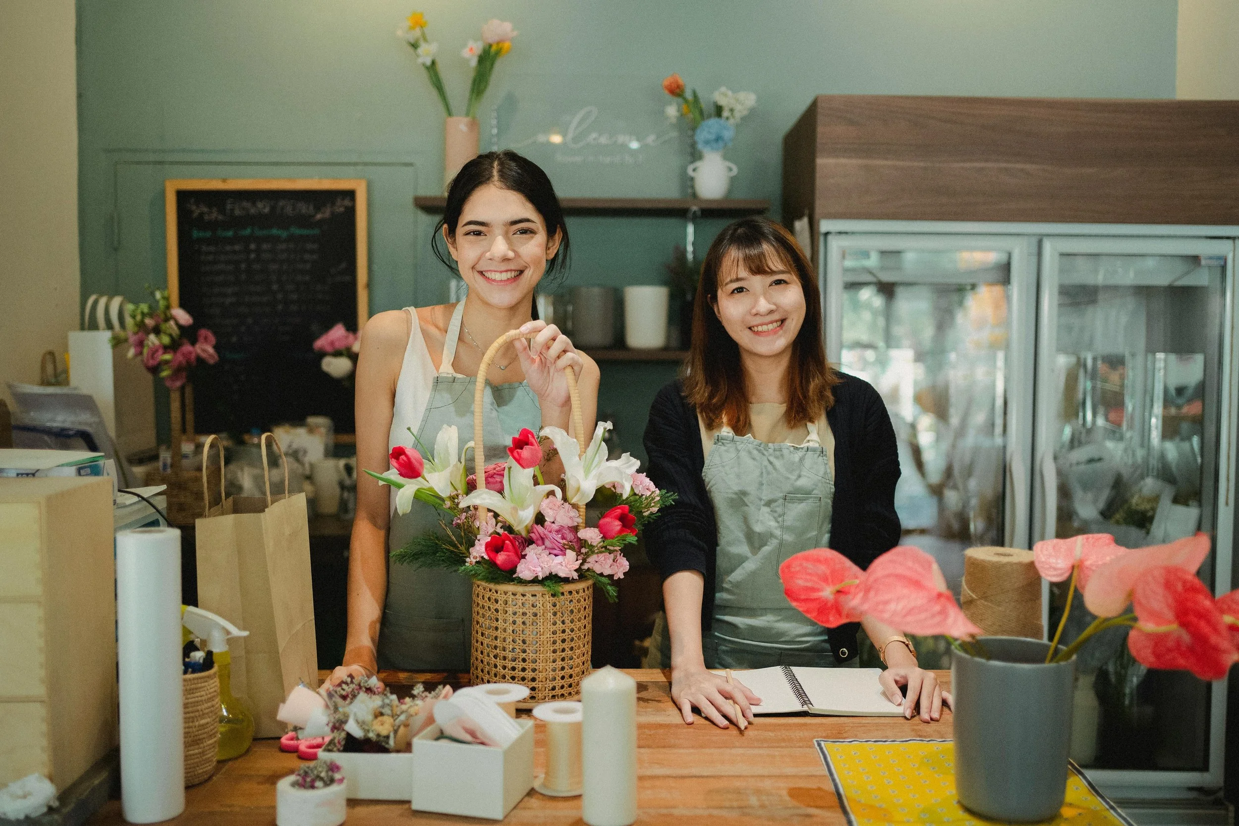 Two women smiling behind a floral display and arranging items at a flower shop. One holding a basket of pink and white flowers, the other with an open notebook, surrounded by flowers, ribbons, and supplies.