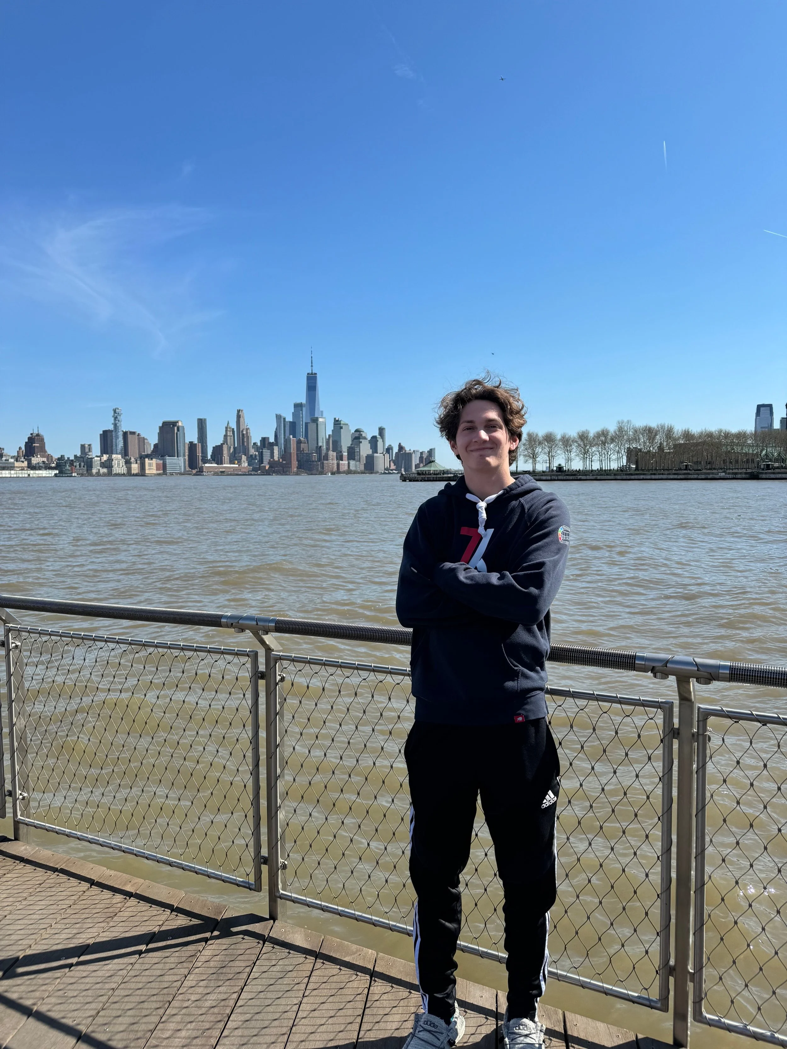 A young man standing with arms crossed on a wooden pier, wearing a black hoodie and black Adidas track pants, with the New York City skyline, including the One World Trade Center, in the background on a clear, sunny day.