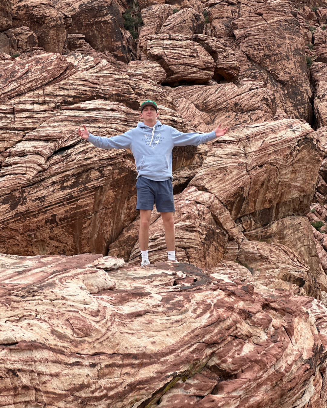 A young man standing with arms outstretched on red and pink layered rocks in a desert landscape.