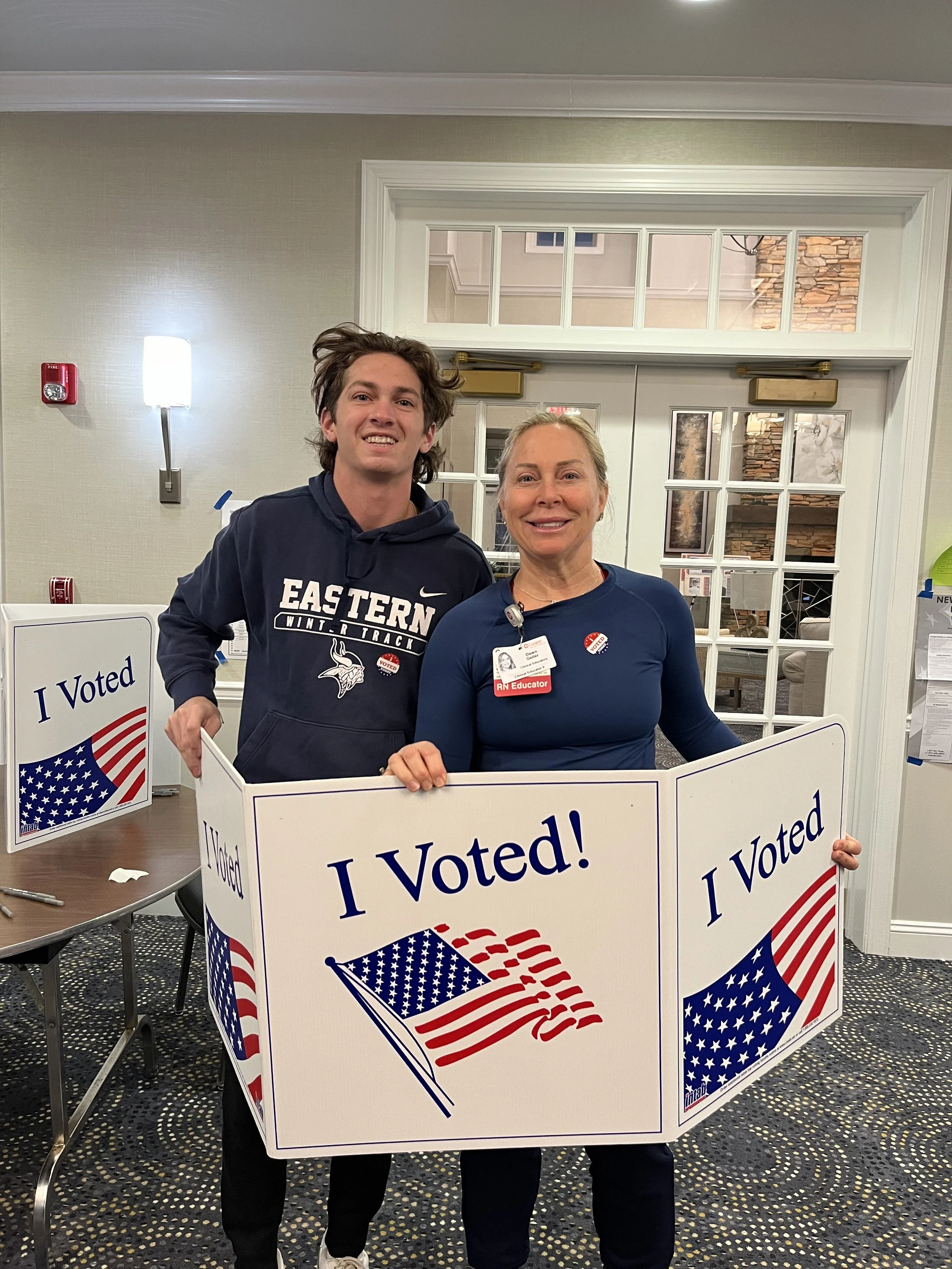 Two people holding a large sign that says "I Voted!" with an American flag and text, both smiling, inside a room near a door and a table with smaller "I Voted" signs.