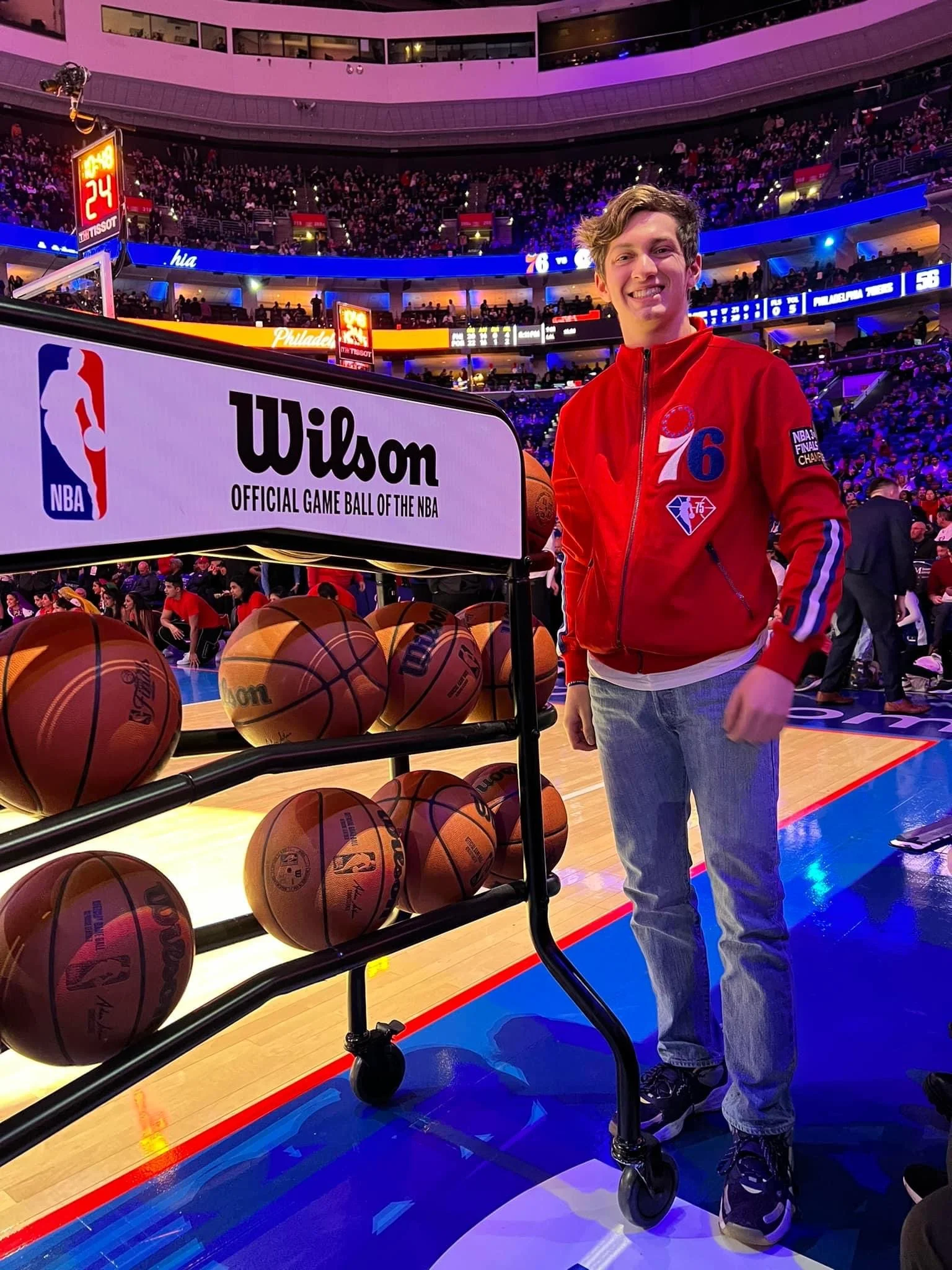 A young man wearing a red Philadelphia 76ers jacket and jeans standing next to a cart filled with basketballs on a basketball court at a game arena.
