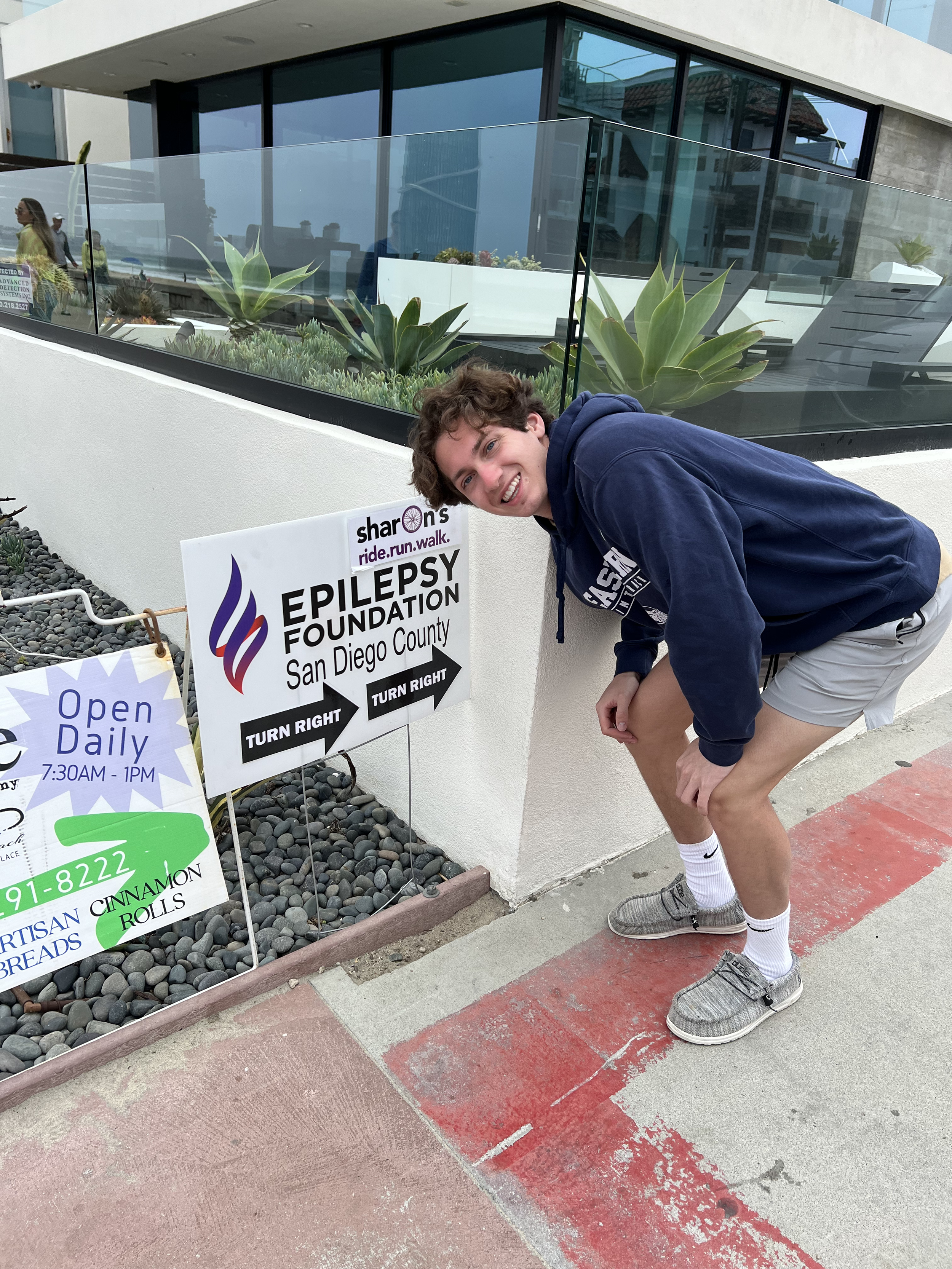 A young man in athletic attire crouches smiling next to signs for the Epilepsy Foundation in San Diego County, with one sign pointing to the right and the other to the left, outside a modern building with large glass windows and desert plants.