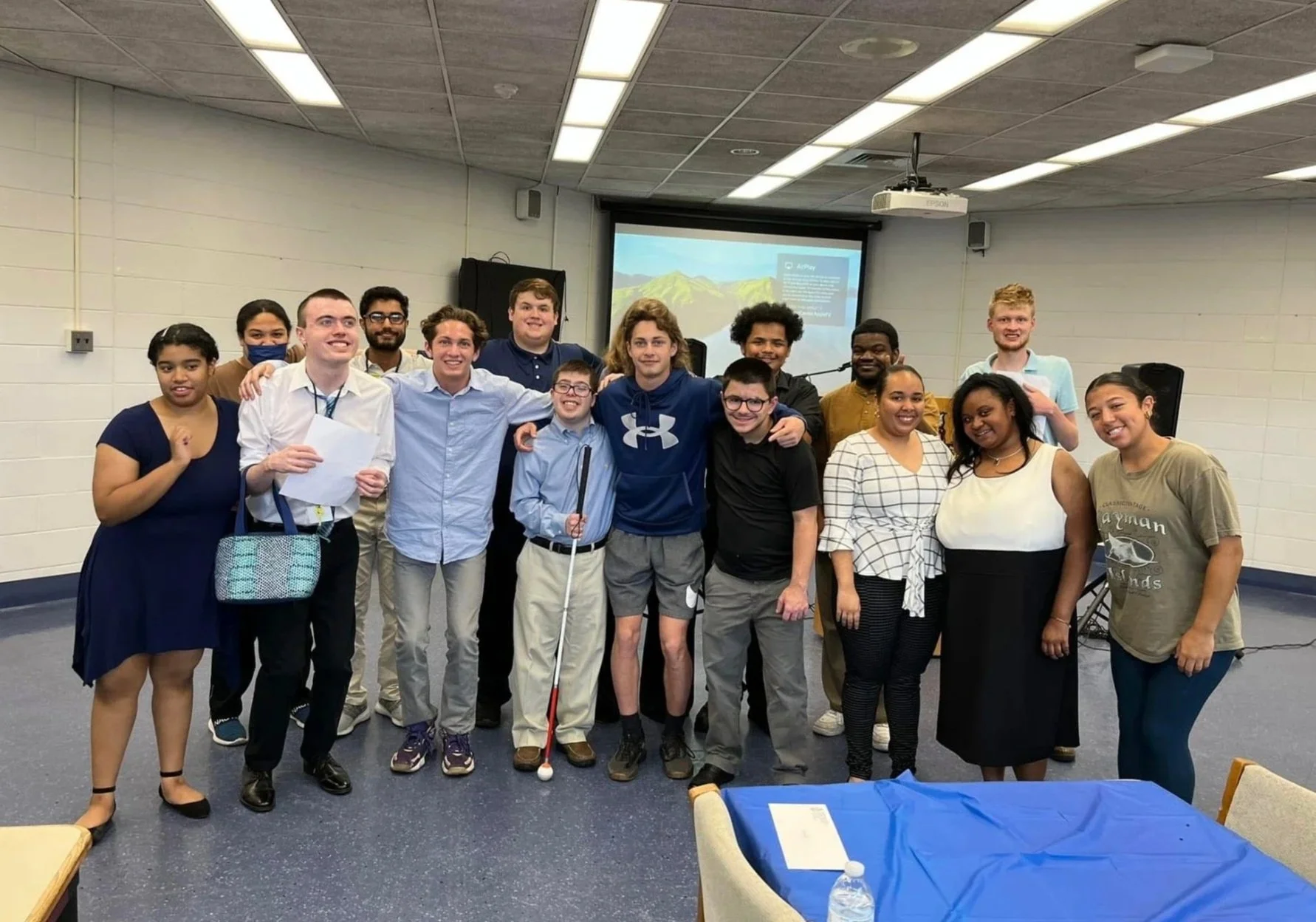 A diverse group of 16 young adults and teens smiling and posing together in a classroom or conference room, with a projector screen and speakers in the background.