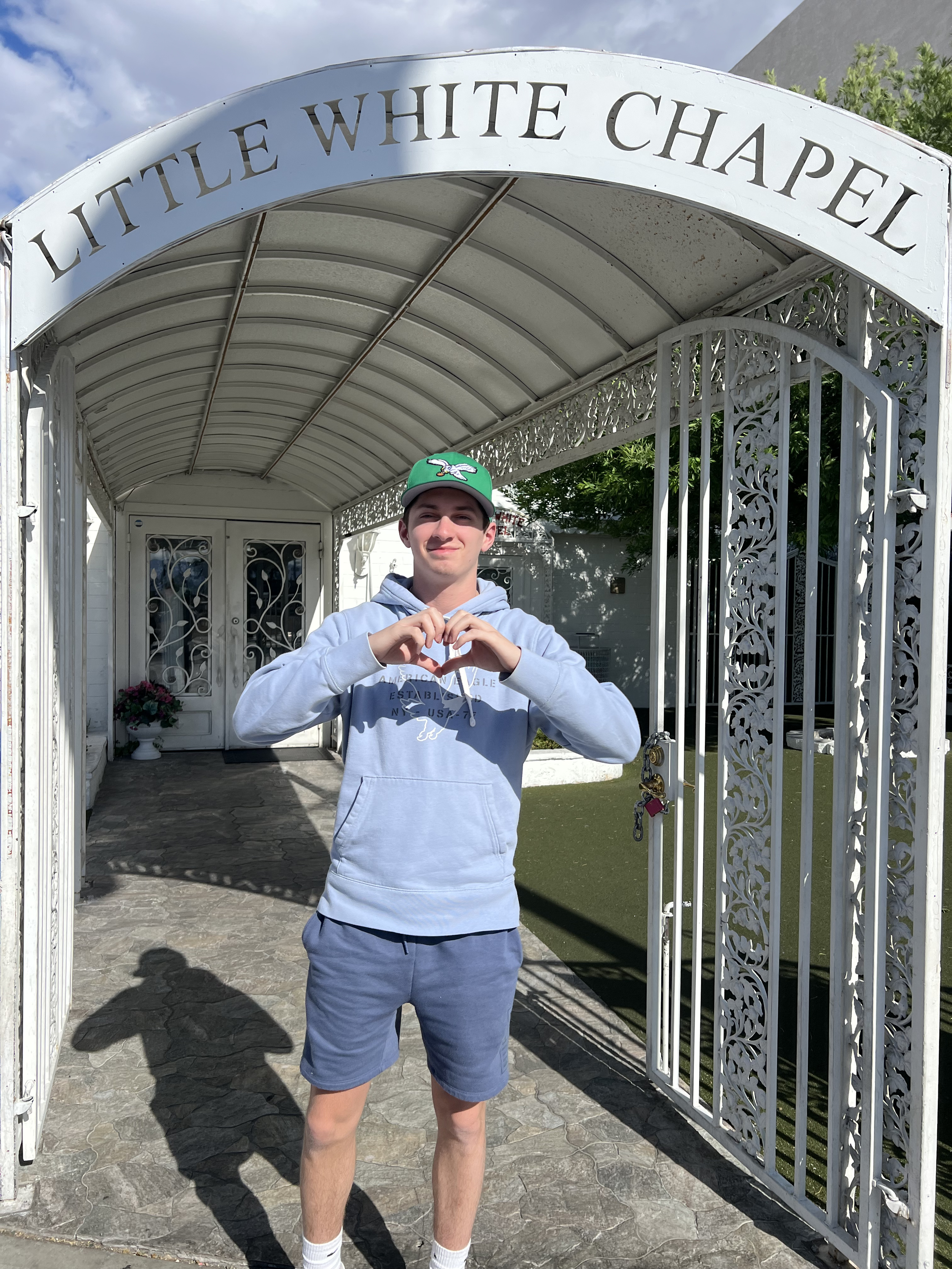 A young man standing under the archway entrance to Little White Chapel, making a heart shape with his hands.